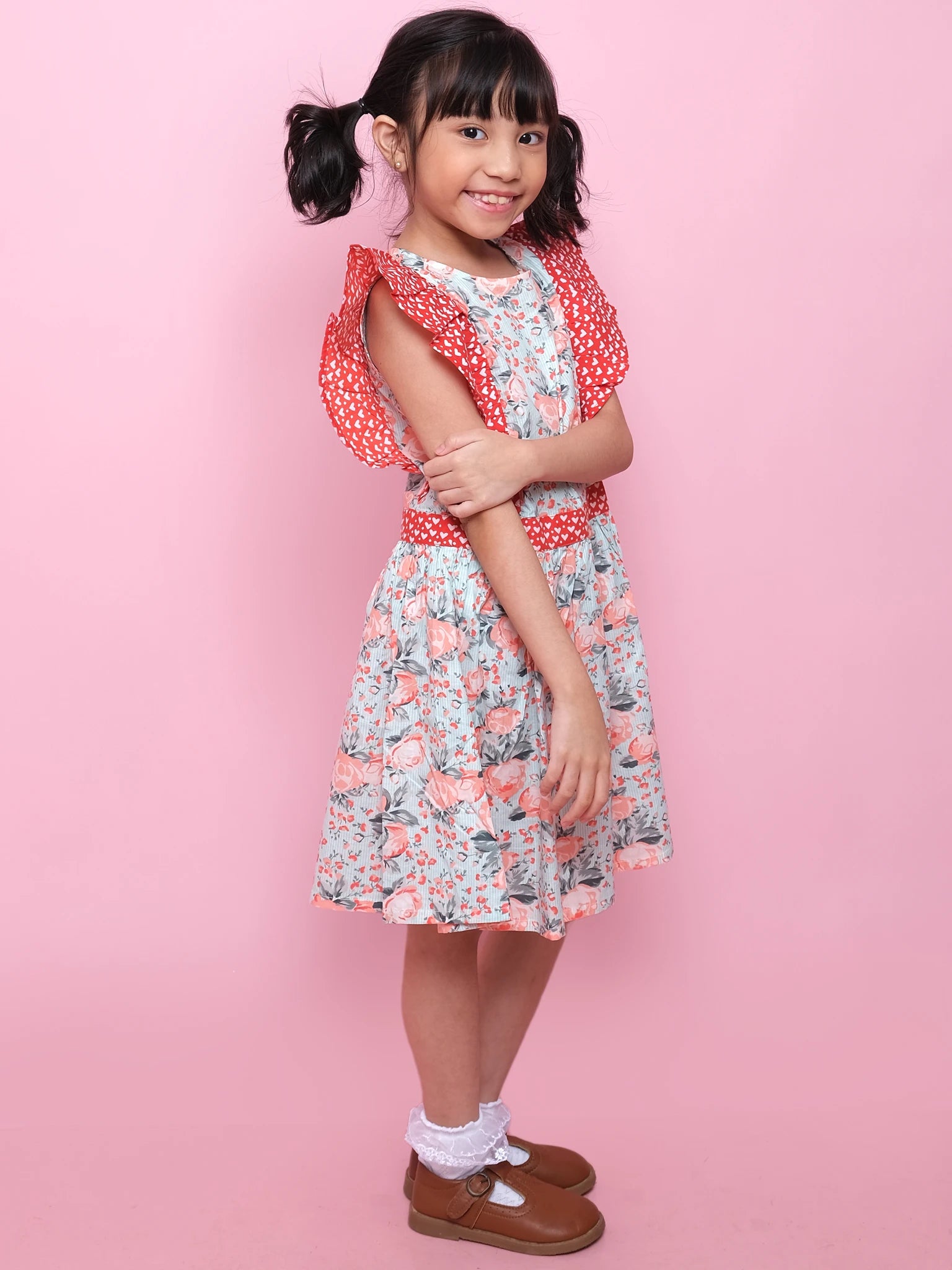 A young girl stands sideways, smiling with arms crossed, wearing the Vintage Roses Jayne Dress by Vintage Roses, paired with white socks and brown shoes, set against a pink background.