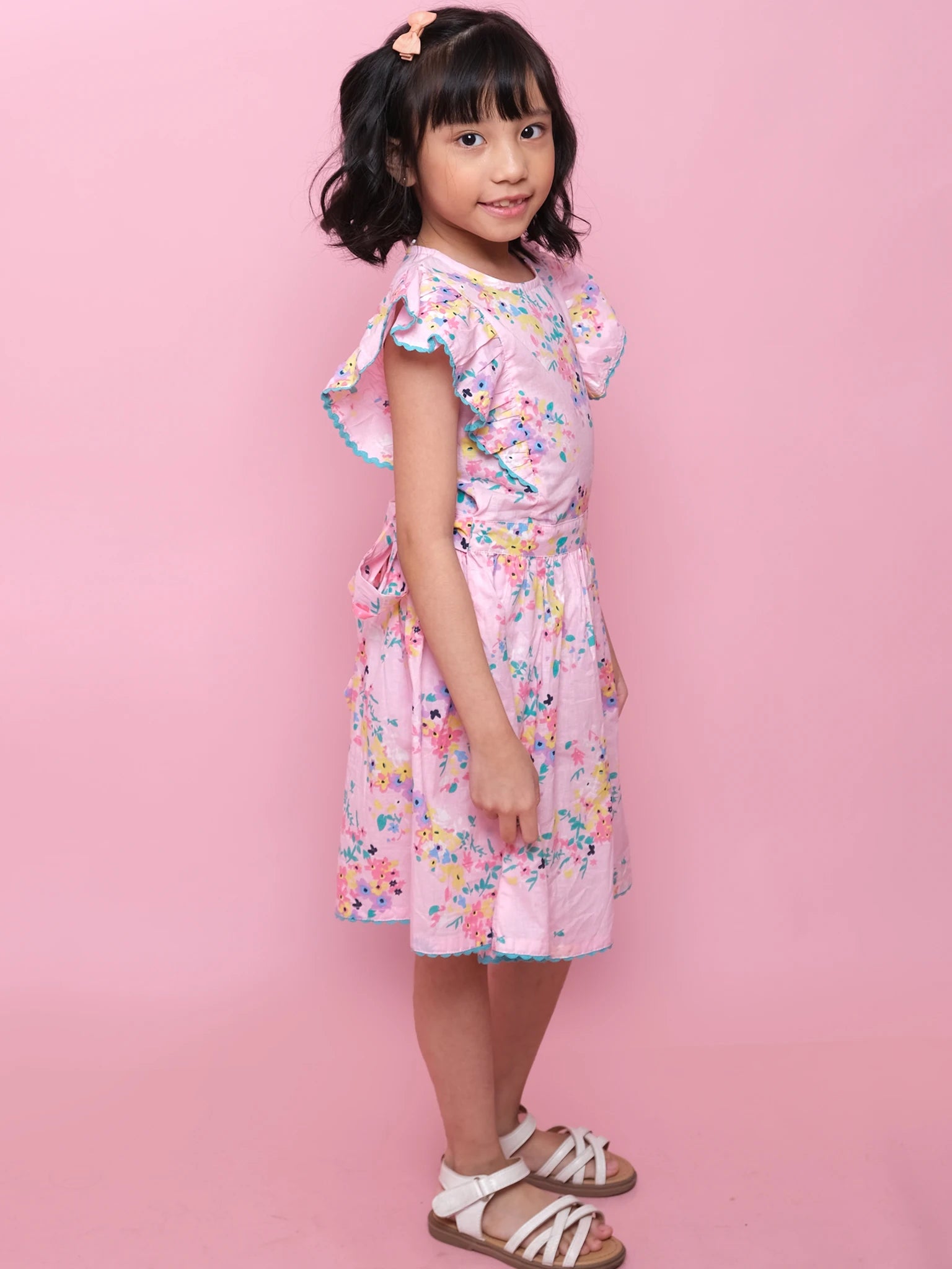 A young girl with short black hair wears the Confetti Floral Lacy Dress by Confetti Floral and white sandals, standing sideways against a pink background.