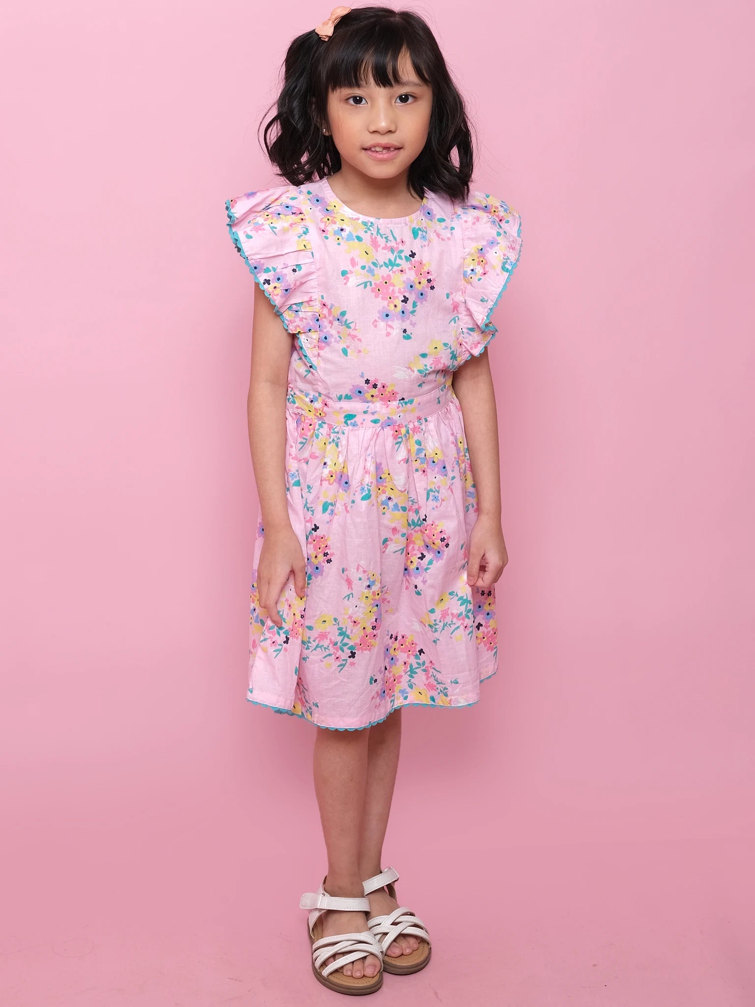 A young girl stands against a pink background in the Confetti Floral Lacy Dress by Confetti Floral— a light pink summer dress with colorful floral patterns, ruffled sleeves, and paired with white sandals.