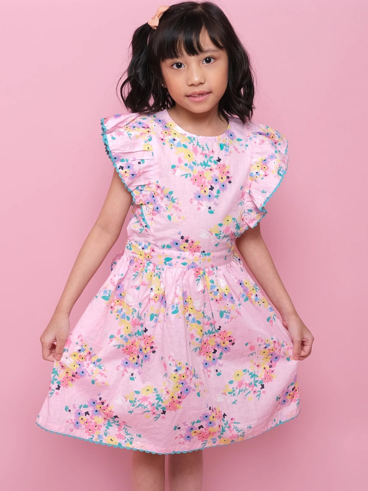 A young girl poses against a pink background, wearing the Confetti Floral Lacy Dress by Confetti Floral—a charming girls’ summer dress with ruffled sleeves and soft floral print, paired with white sandals.