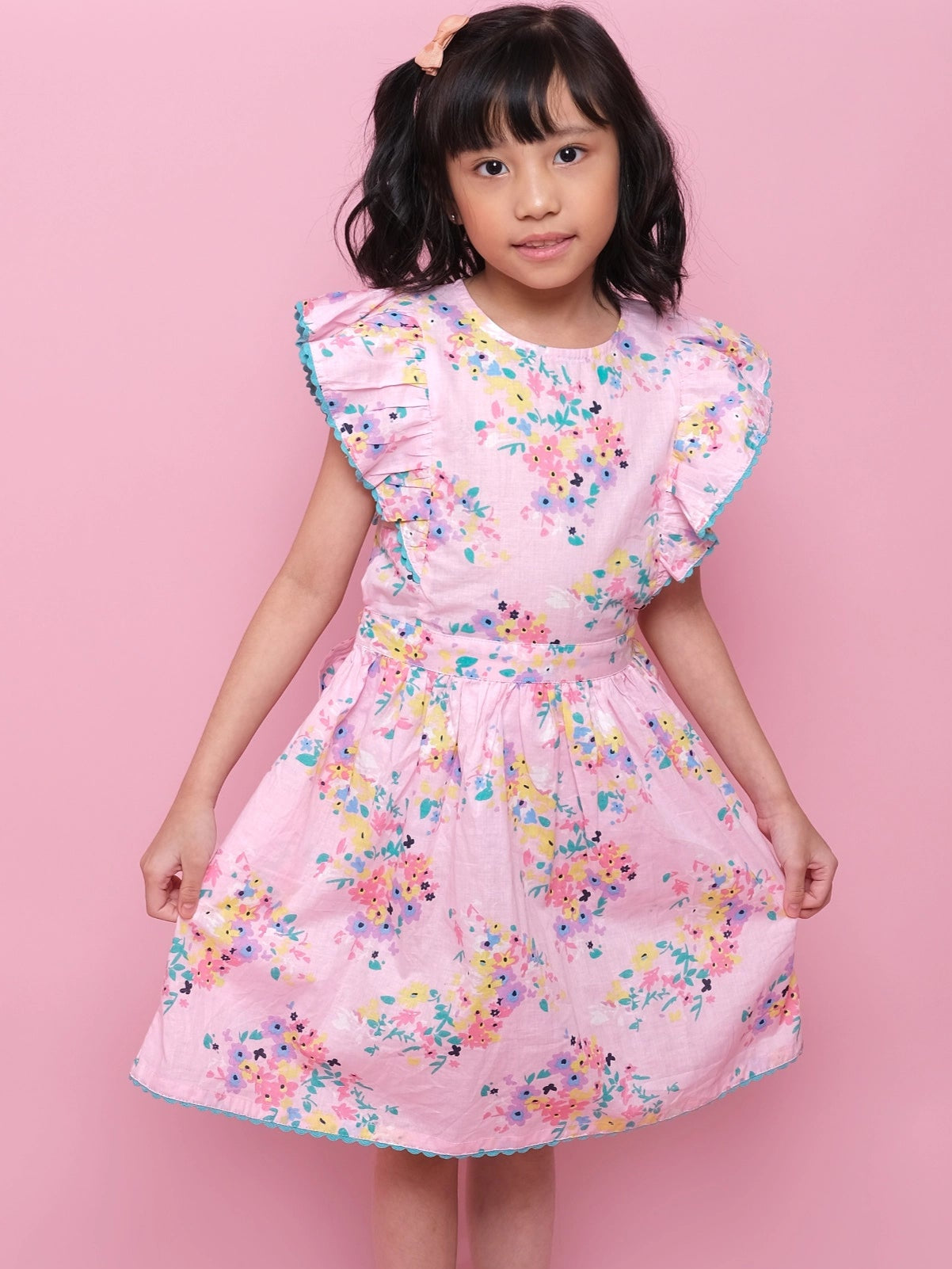 A young girl models the Confetti Floral Lacy Dress by Confetti Floral, featuring pink floral lace, ruffled sleeves, and a charming design, as she poses holding the sides of her dress against a solid pink background.