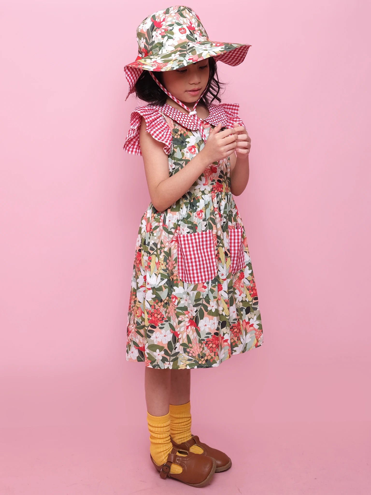A young girl wears the Aussie Bush Flowers Collared Dress by Aussie Bush Flowers with a matching wide-brim hat, yellow socks, and brown shoes, standing against a pink background.
