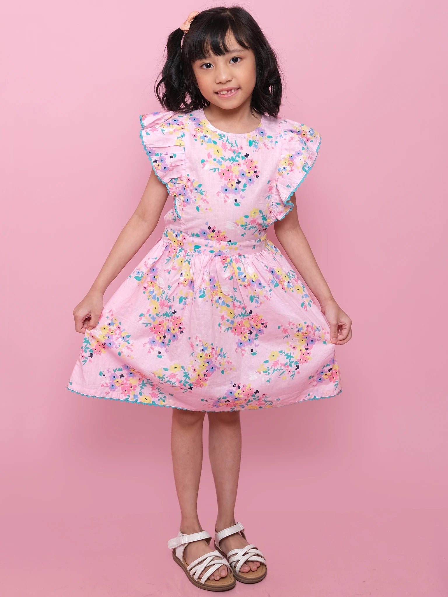 A young girl stands against a pink background wearing the Confetti Floral Lacy Dress by Confetti Floral, featuring lace trim and ruffled sleeves, paired with white sandals and holding the sides of her dress.