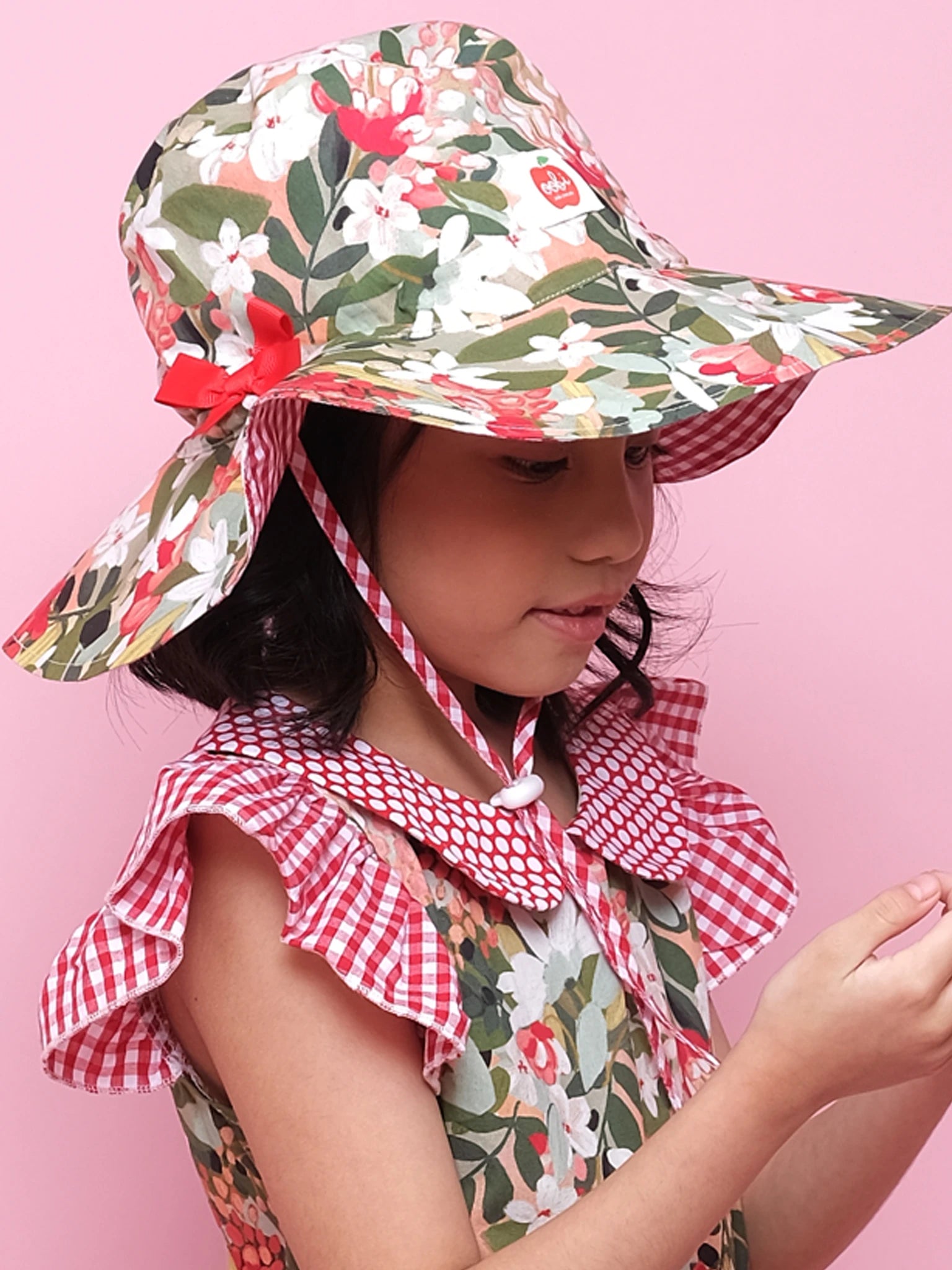 A young girl wearing an Aussie Bush Flowers Ponytail Hat by Aussie Bush Flowers and a matching floral and gingham dress stands against a pink background, looking at something in her hands.