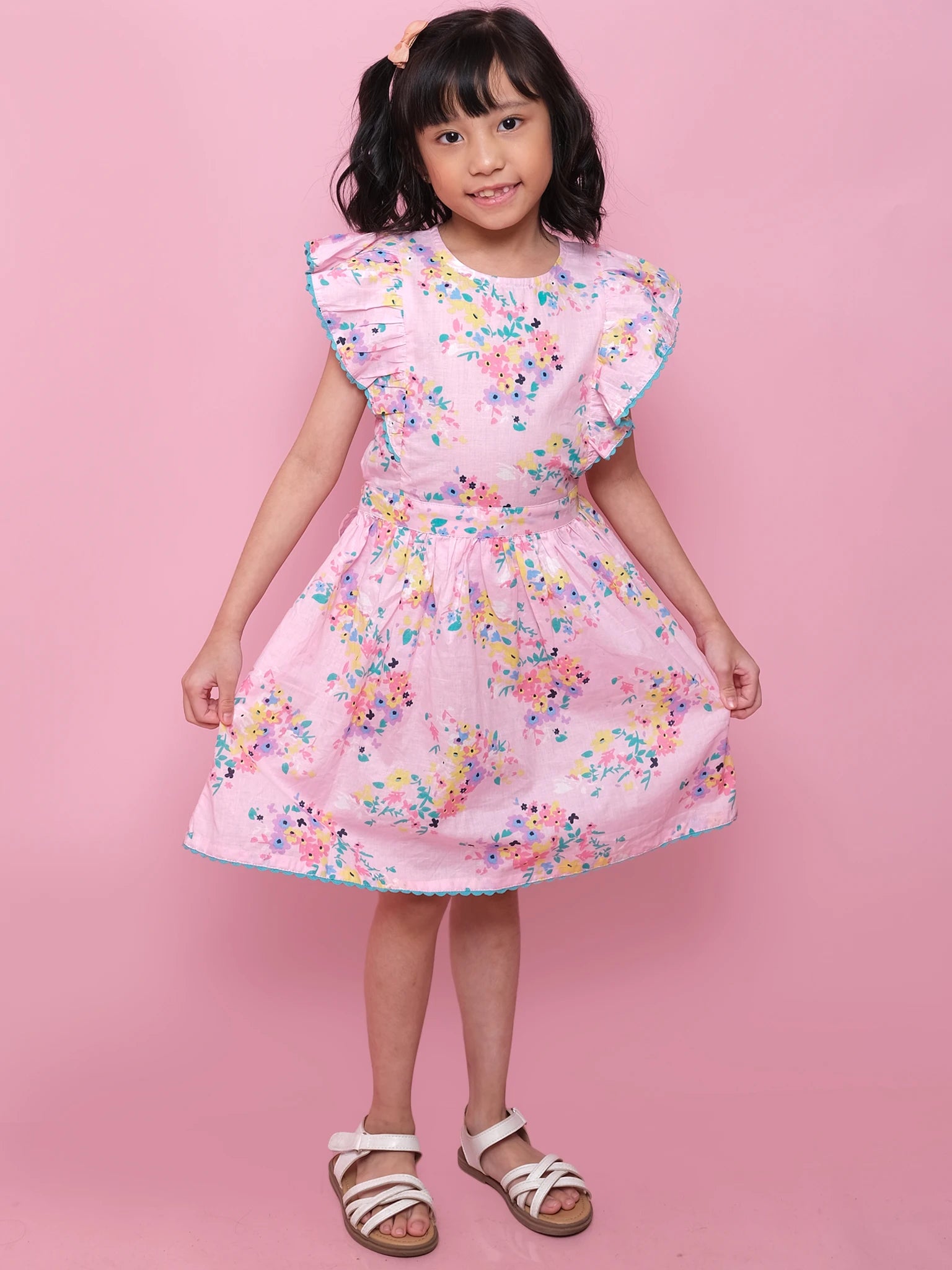 A young girl poses against a pink background wearing the Confetti Floral Lacy Dress by Confetti Floral, featuring lace trim and ruffled sleeves, paired with white sandals as she holds the sides of her dress.