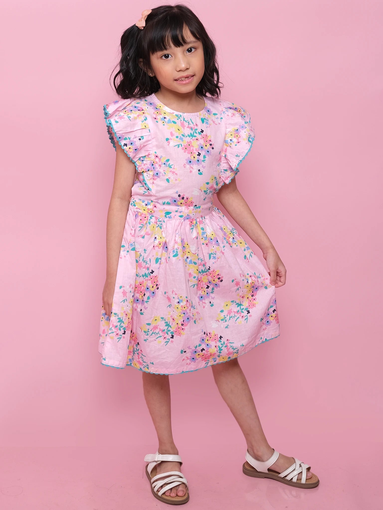 A young girl stands against a pink backdrop wearing the Confetti Floral Lacy Dress by Confetti Floral, featuring ruffled sleeves and lace trim. She completes the look with white sandals and a pink hair clip in her wavy black hair.