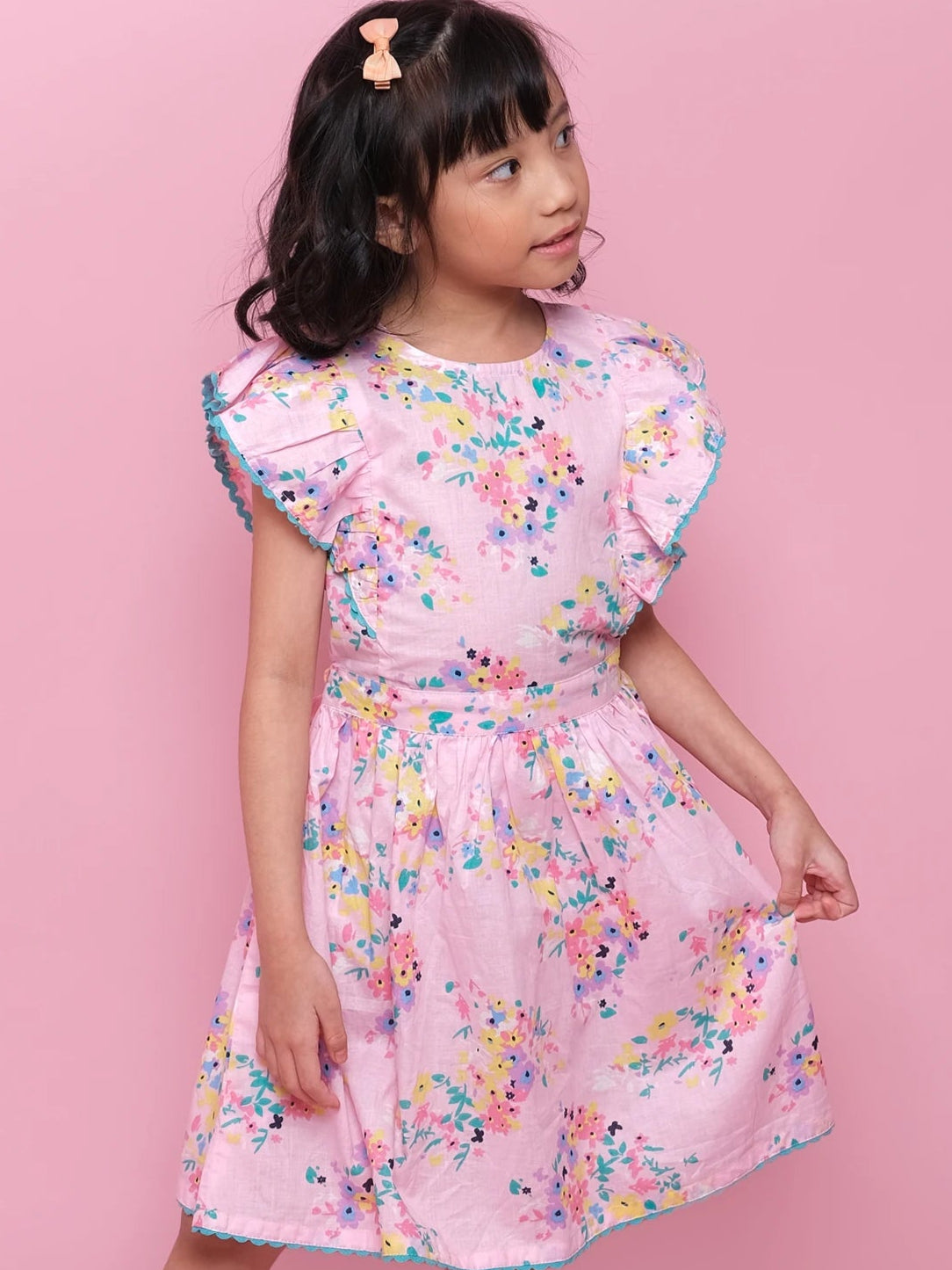 A young girl wearing the Confetti Floral Lacy Dress by Confetti Floral, featuring a pink floral print with ruffled sleeves, pairs it with white sandals and stands against a solid pink background.