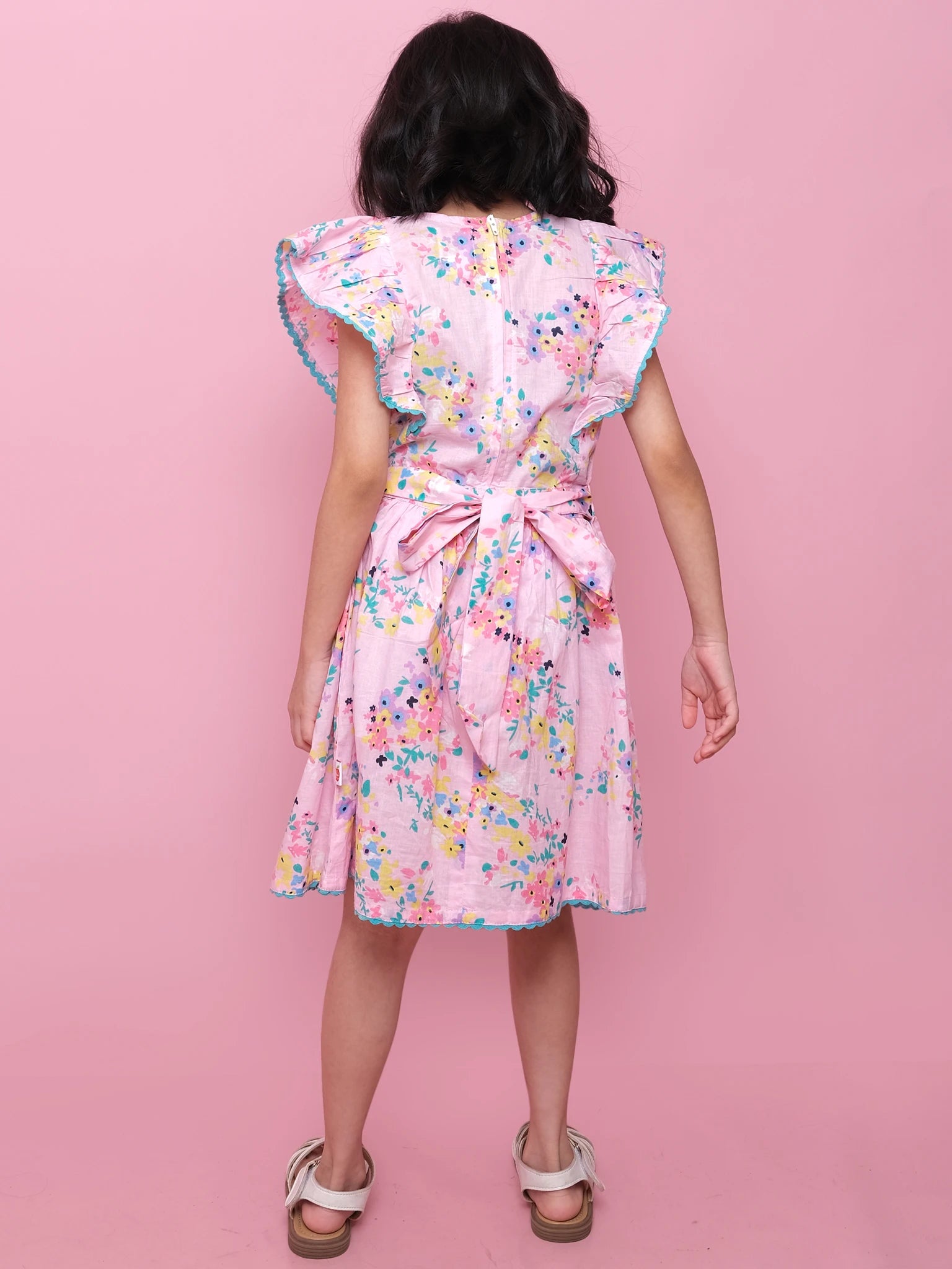A girl, back to camera, wears the Confetti Floral Lacy Dress by Confetti Floral—a pink floral dress with lace trim, ruffled sleeves, a large waist bow, and white sandals—against a pink background.