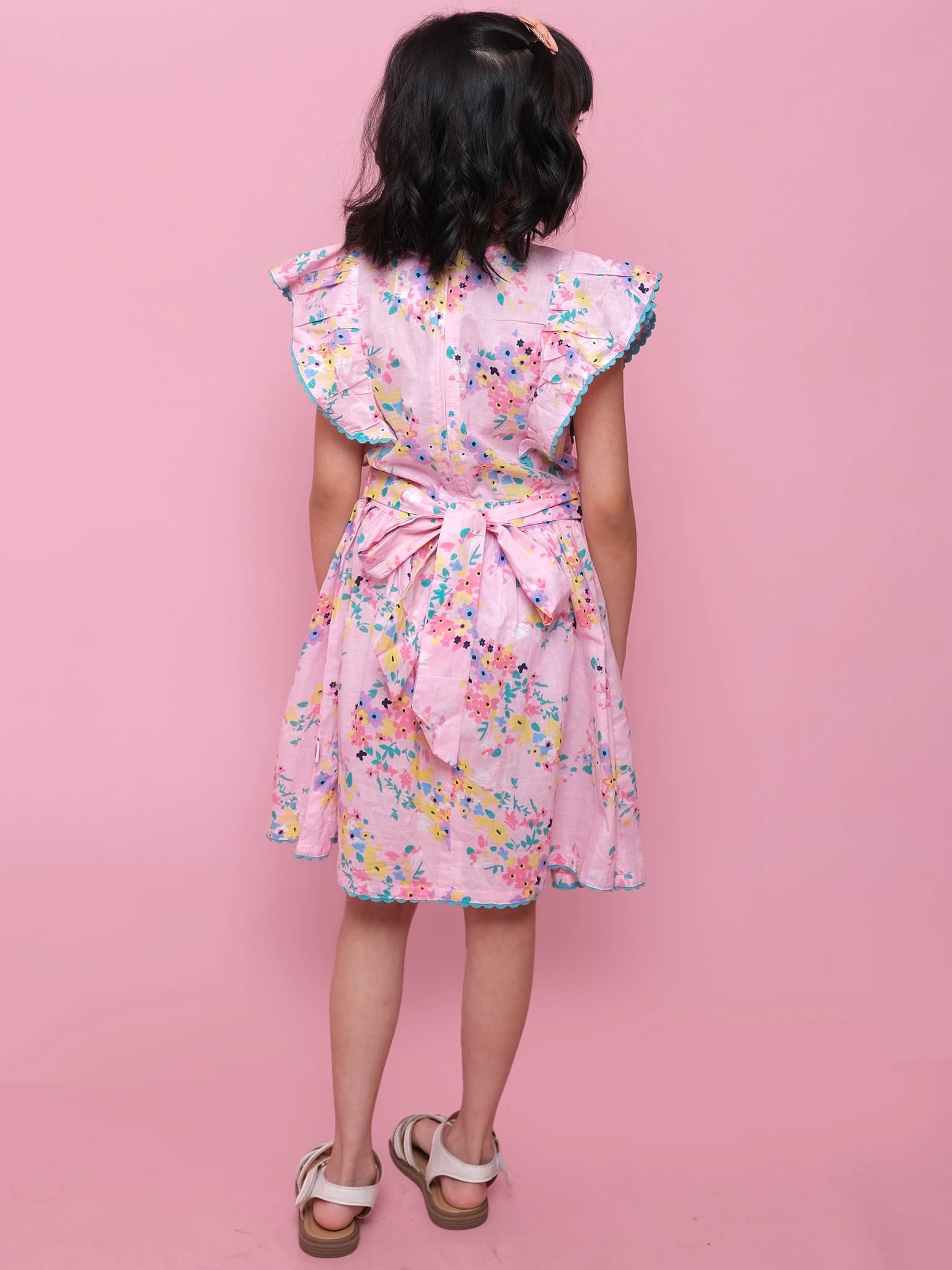 A young girl faces away in the Confetti Floral Lacy Dress by Confetti Floral, featuring pink floral lace, ruffled sleeves, a large back bow, and white sandals against a solid pink background.