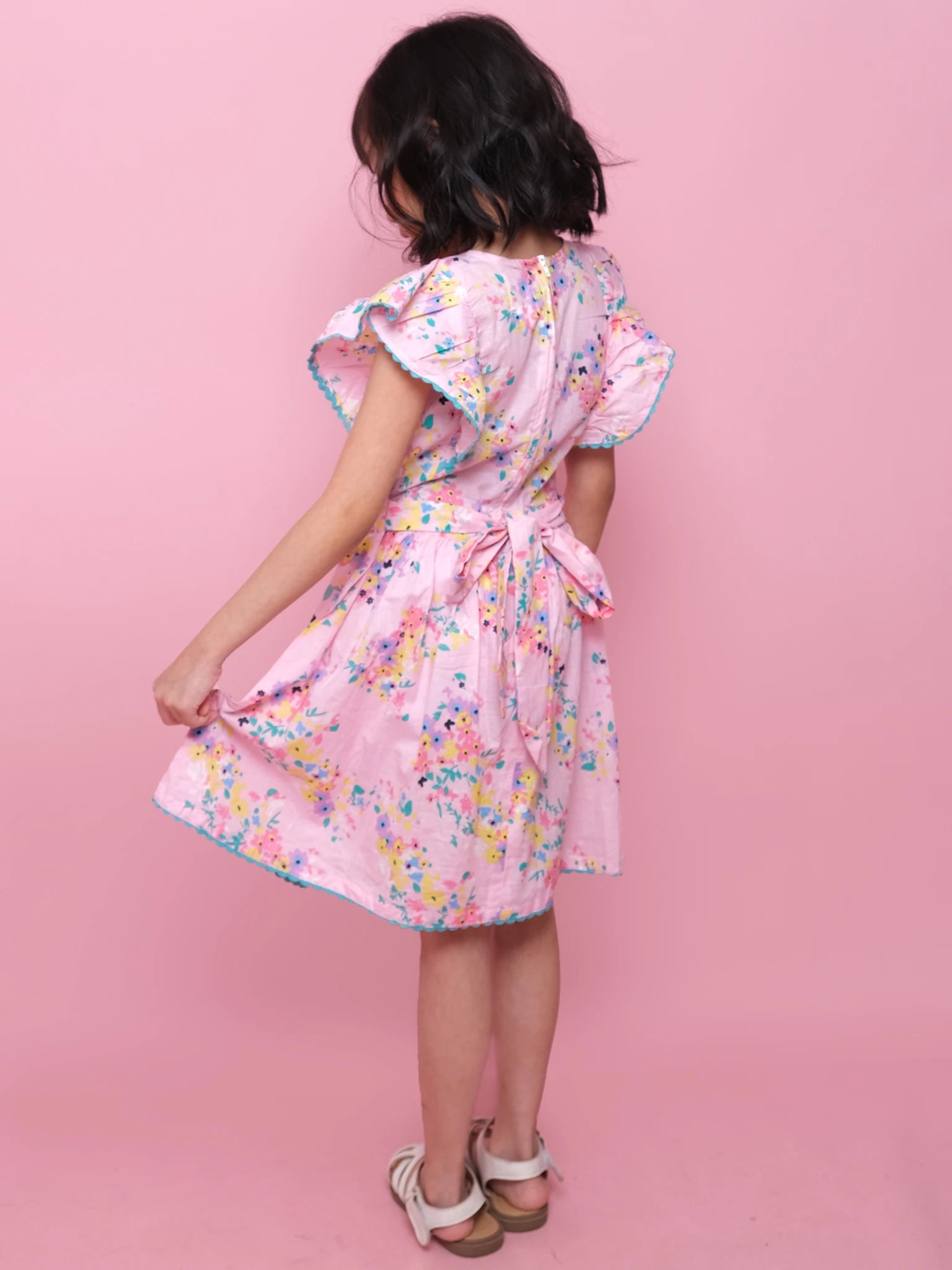 A child models the Confetti Floral Lacy Dress by Confetti Floral, featuring pink floral cotton and a back bow, while standing against a pink background, facing away from the camera.