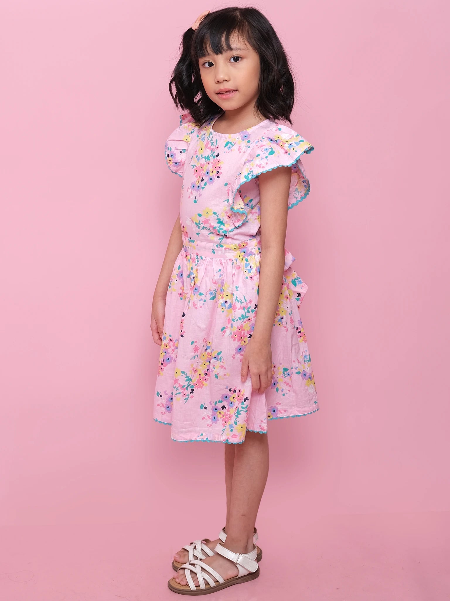 A young girl stands against a pink background wearing the Confetti Floral Lacy Dress by Confetti Floral, a light pink summer dress with lace trim and ruffled sleeves, paired with white sandals.