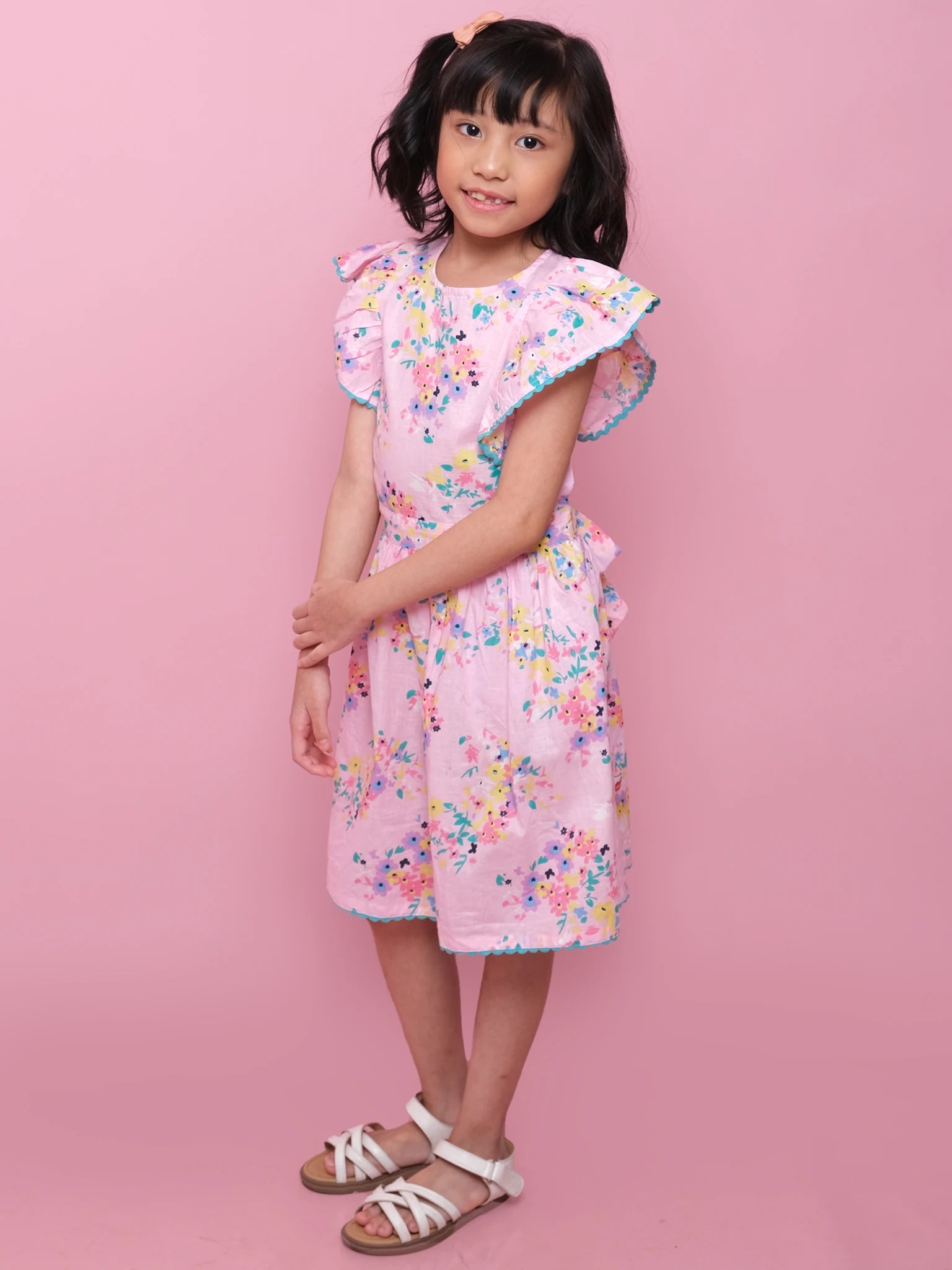 A young girl poses against a pink background, wearing the Confetti Floral Lacy Dress with ruffled sleeves and lace trim from Confetti Floral, paired with white sandals. The perfect summer dress for any occasion.