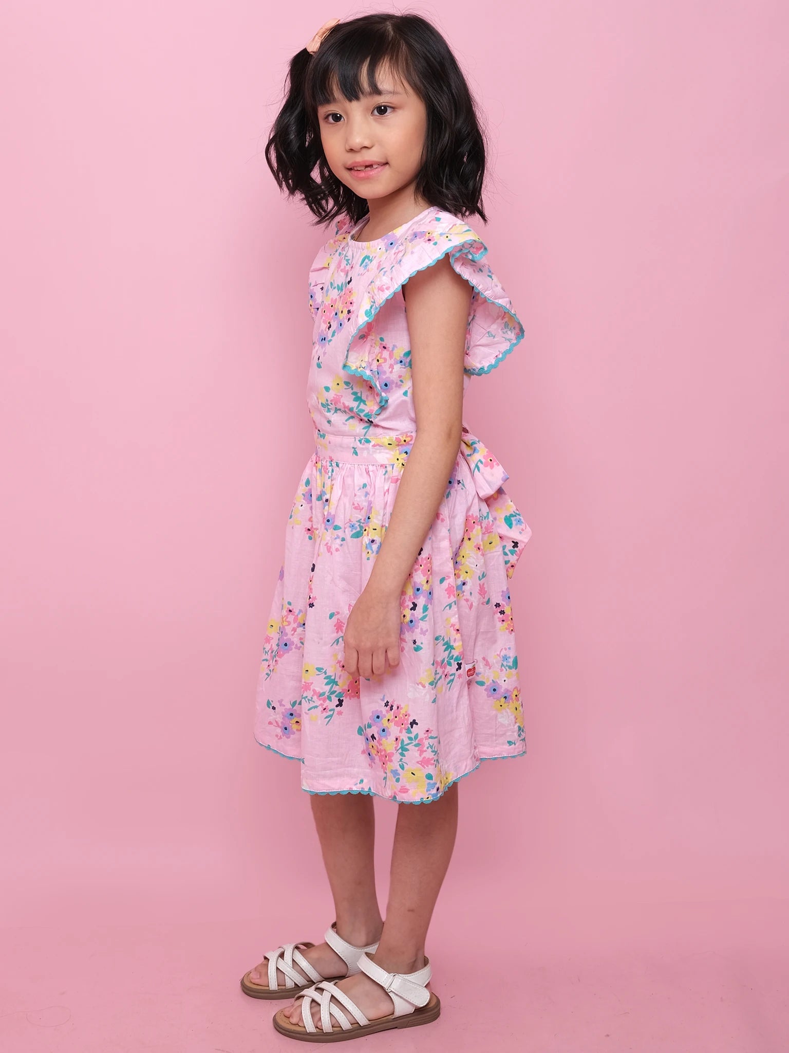 A young girl with shoulder-length dark hair stands sideways against a pink background, wearing the Confetti Floral Lacy Dress and white sandals.