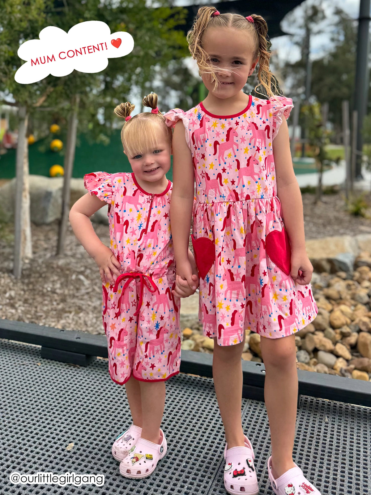 Two young girls standing outdoors on a mesh platform, holding hands and smiling at the camera. Both wear matching pink outfits featuring a pattern of pink and red horses, yellow stars, and blue sparkles.