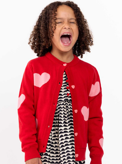 A young girl in a Maxine Red with Pink Hearts Cardigan by Knitwear yawns widely, standing against a white background.