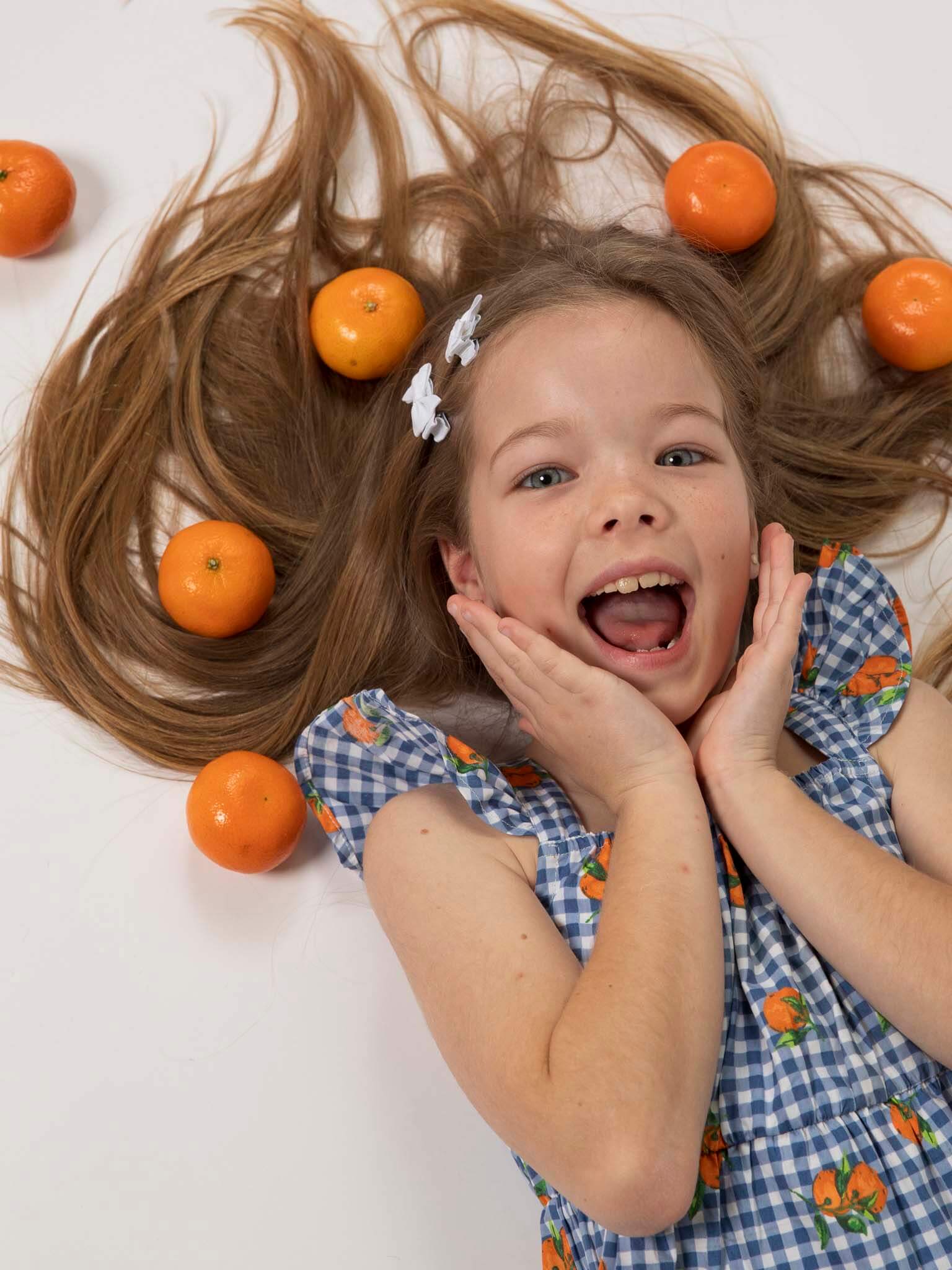 A young girl with long brown hair wears the Amalfi Oranges Holiday Dress by Amalfi Oranges, lying on the floor with oranges in her hair and smiling brightly.