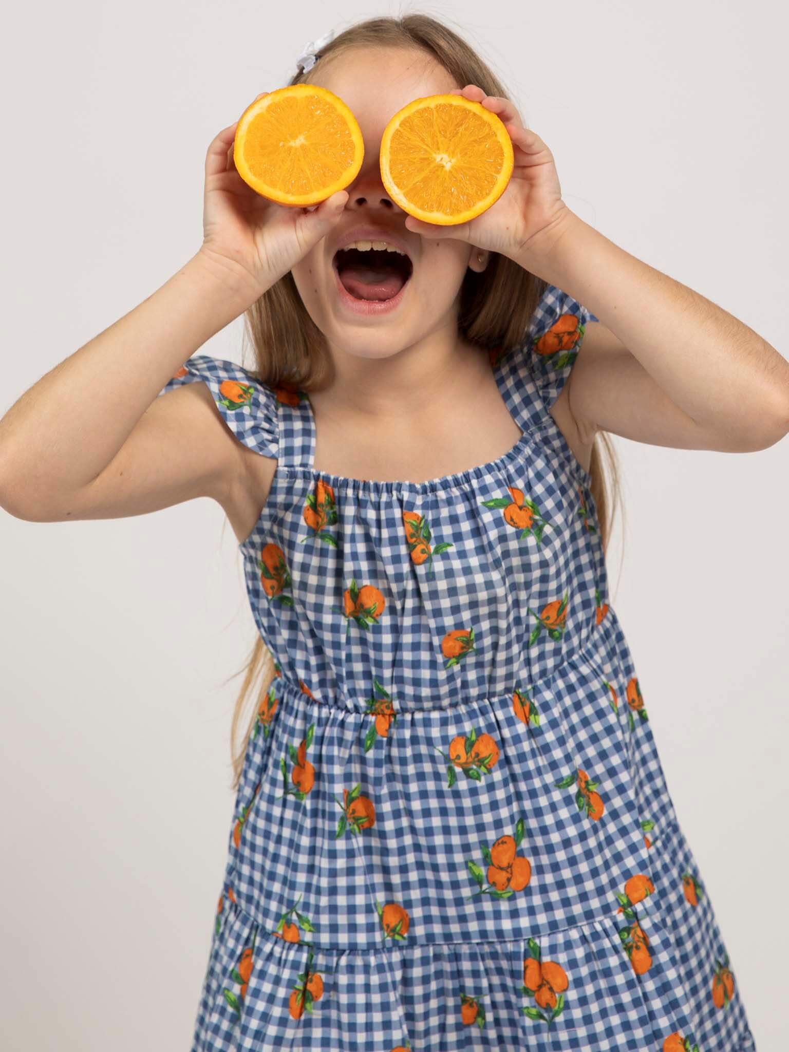 A girl in the Amalfi Oranges Holiday Dress by Amalfi Oranges, featuring blue and white checks with orange prints, holds two orange halves over her eyes and smiles widely.