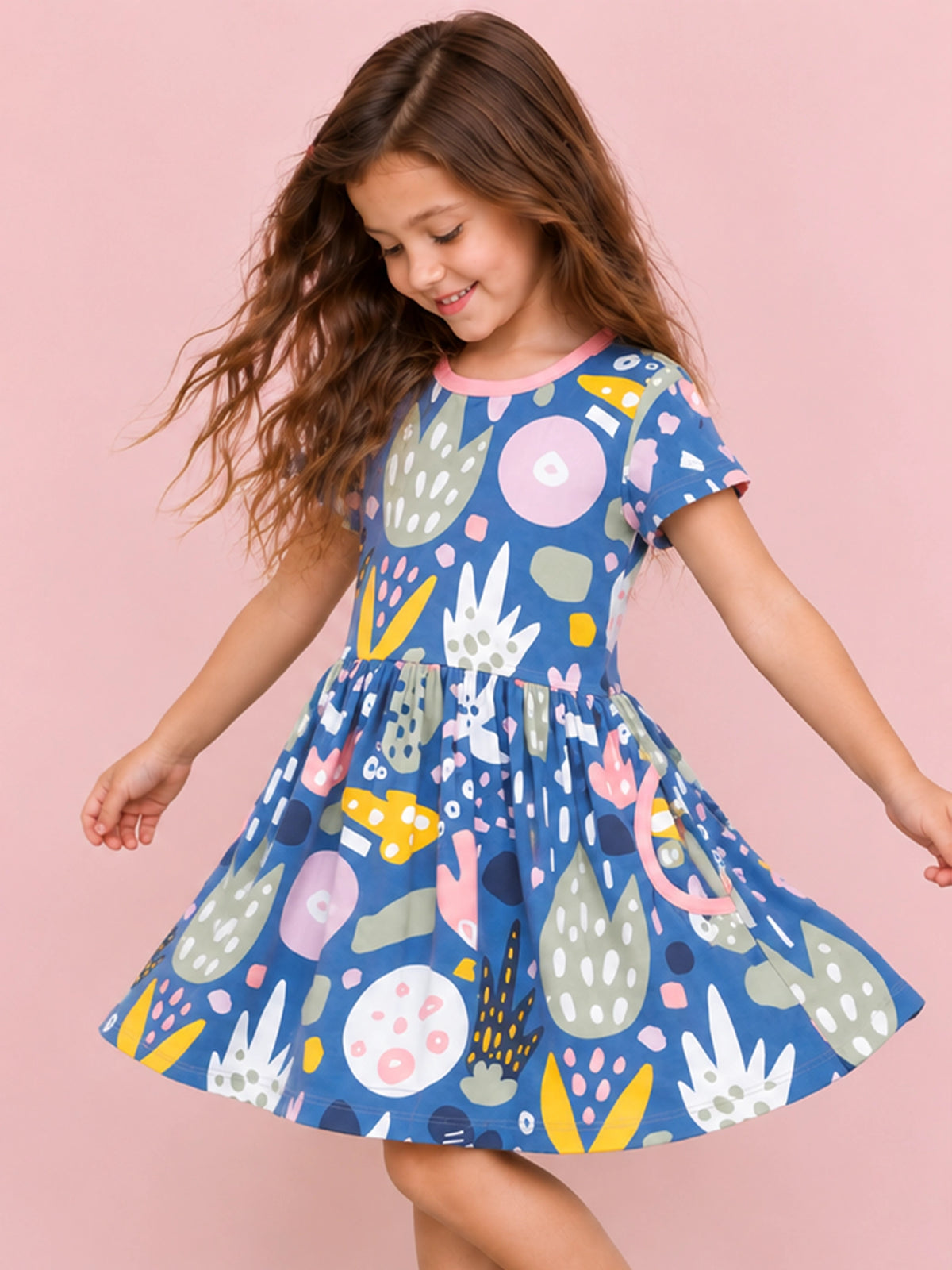 A young girl with long brown hair is wearing a short-sleeve blue dress with a colorful abstract pattern, posing and twirling against a solid pink background.