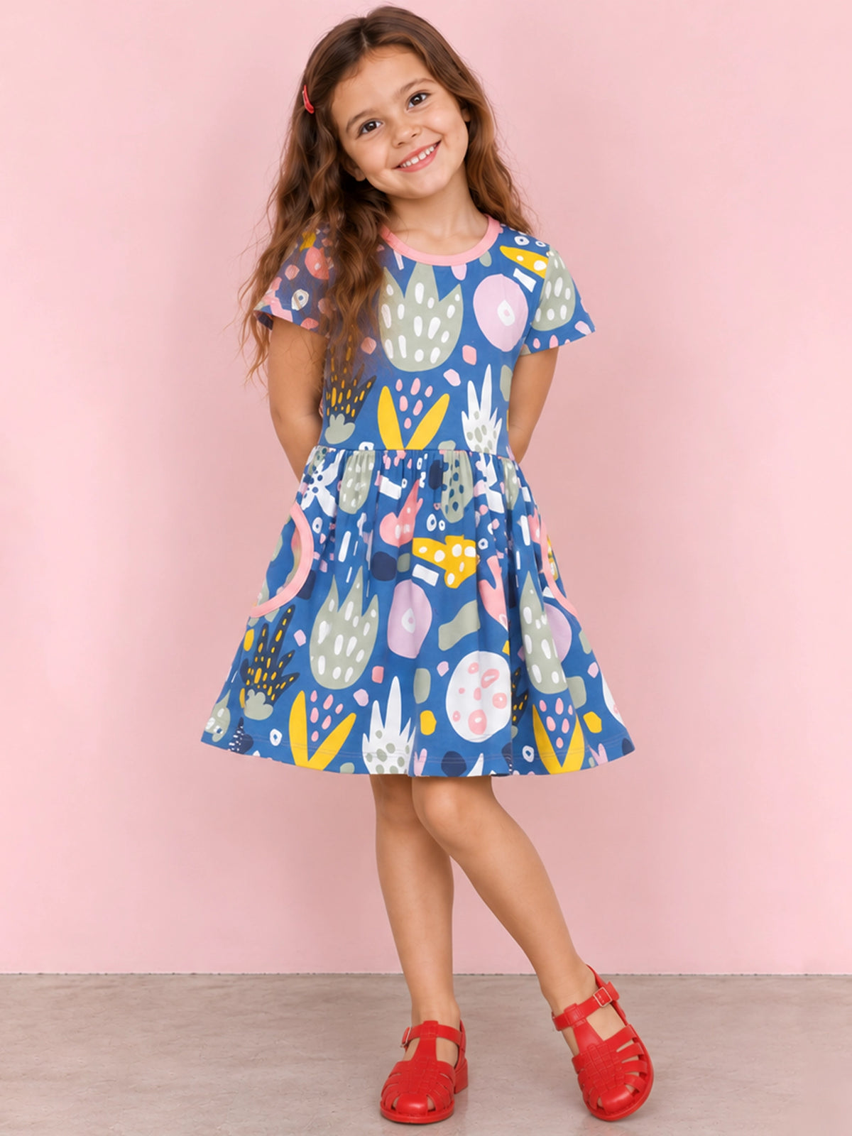 A young girl with long brown hair is wearing a short-sleeve blue dress with a colorful abstract pattern and red sandals, standing against a solid pink background.