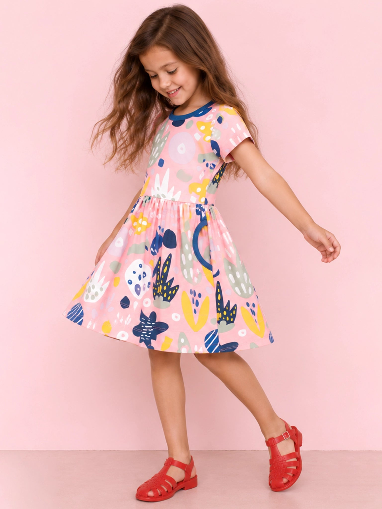 A young girl with long brown hair is wearing a pink short-sleeve dress with a colorful abstract pattern and red sandals, posing against a solid pink background.