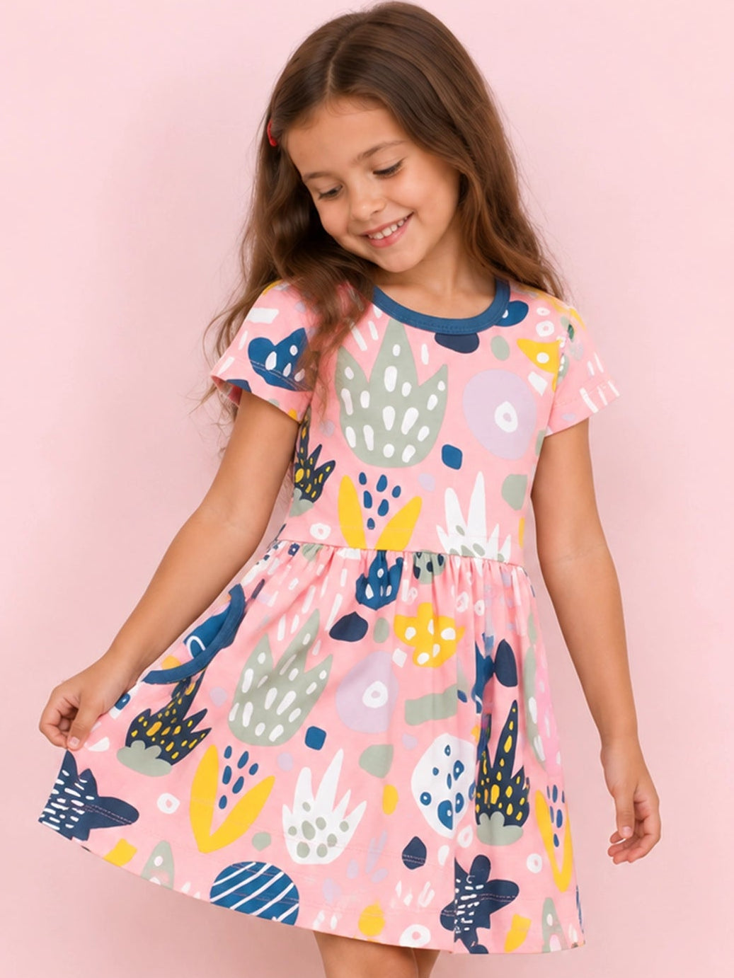 A young girl with long brown hair is wearing a pink short-sleeve dress with a colorful abstract pattern, standing against a solid pink background. She is wearing red shoes.