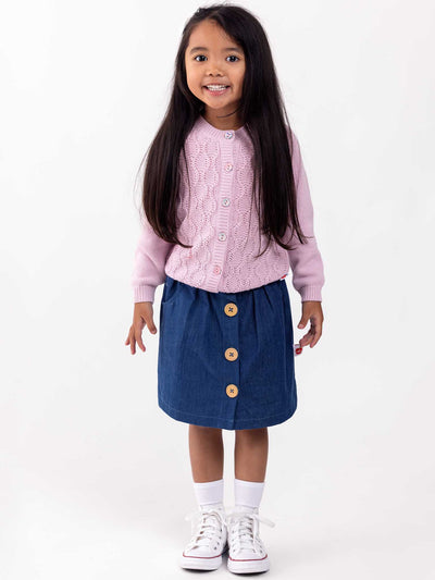 A young girl standing and smiling, wearing a pink sweater, a Red Rainbow Dark Denim Button Skirt, white socks, and white sneakers on a plain background.