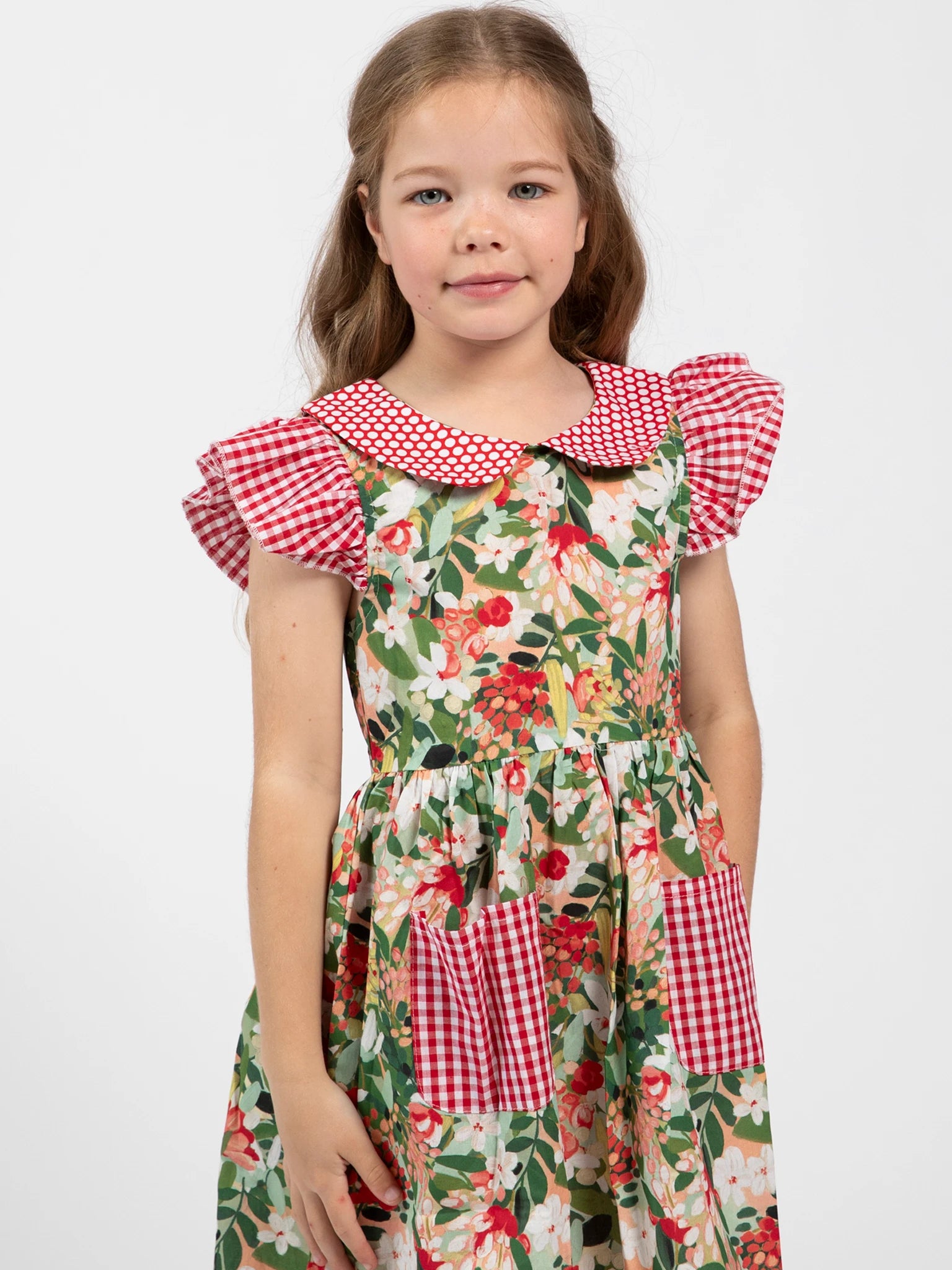 A young girl with long brown hair models the Aussie Bush Flowers Collared Dress by Aussie Bush Flowers, featuring red and white checkered details and a floral print, made from 100% cotton, against a plain white background.