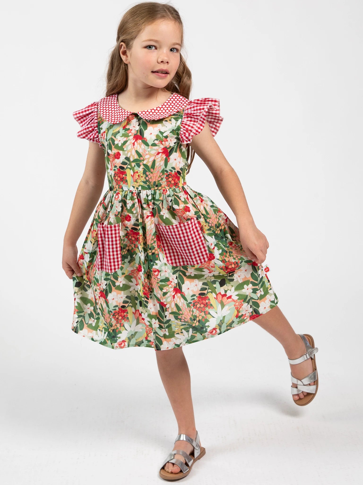 A young girl models the Aussie Bush Flowers Collared Dress by Aussie Bush Flowers, featuring a 100% cotton floral print with red gingham sleeves, collar, and pockets, against a plain white background.
