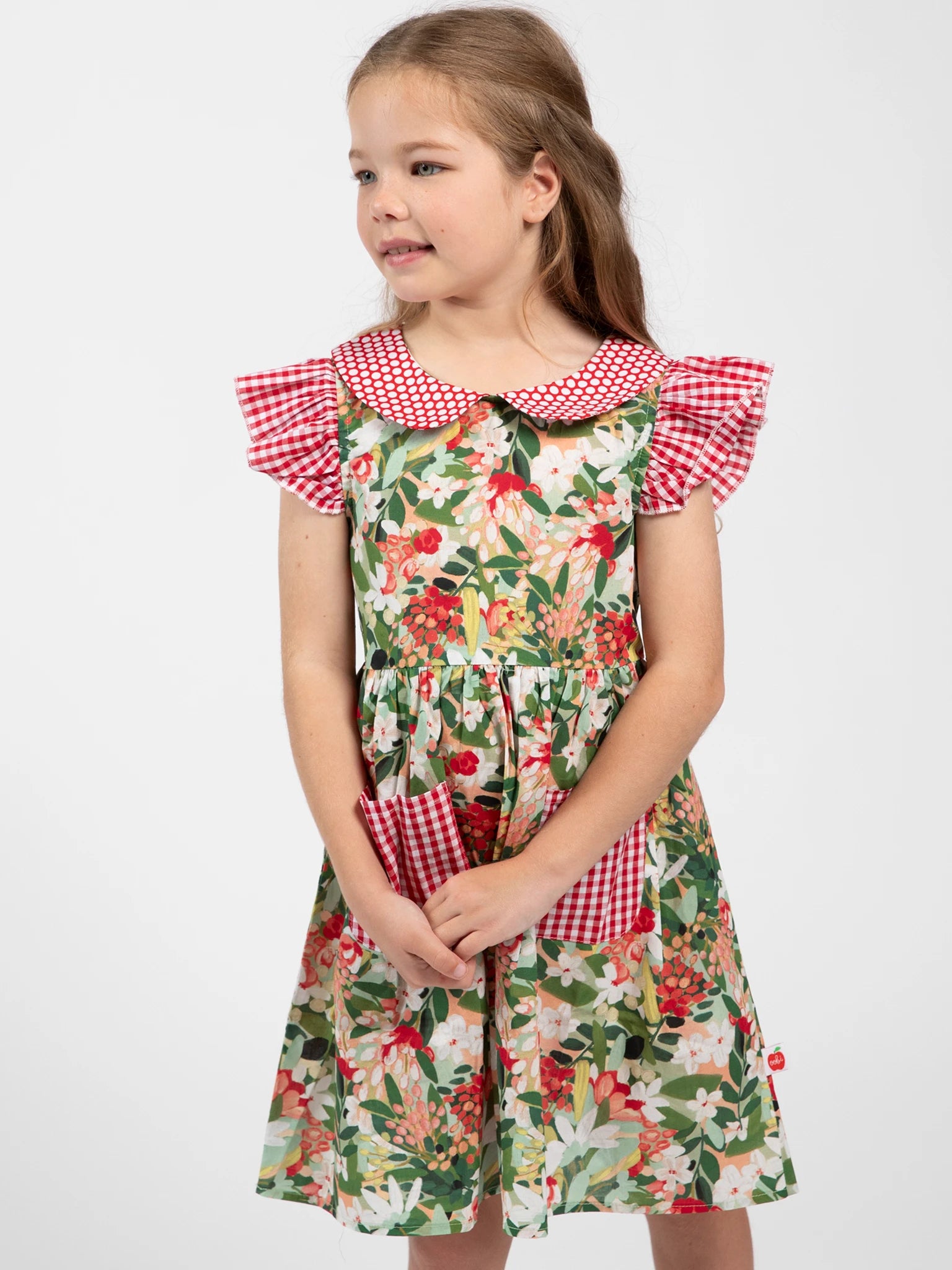 A young girl wears the Aussie Bush Flowers Collared Dress by Aussie Bush Flowers, featuring 100% cotton fabric, floral print, and red gingham collar and sleeves, standing against a plain white background.