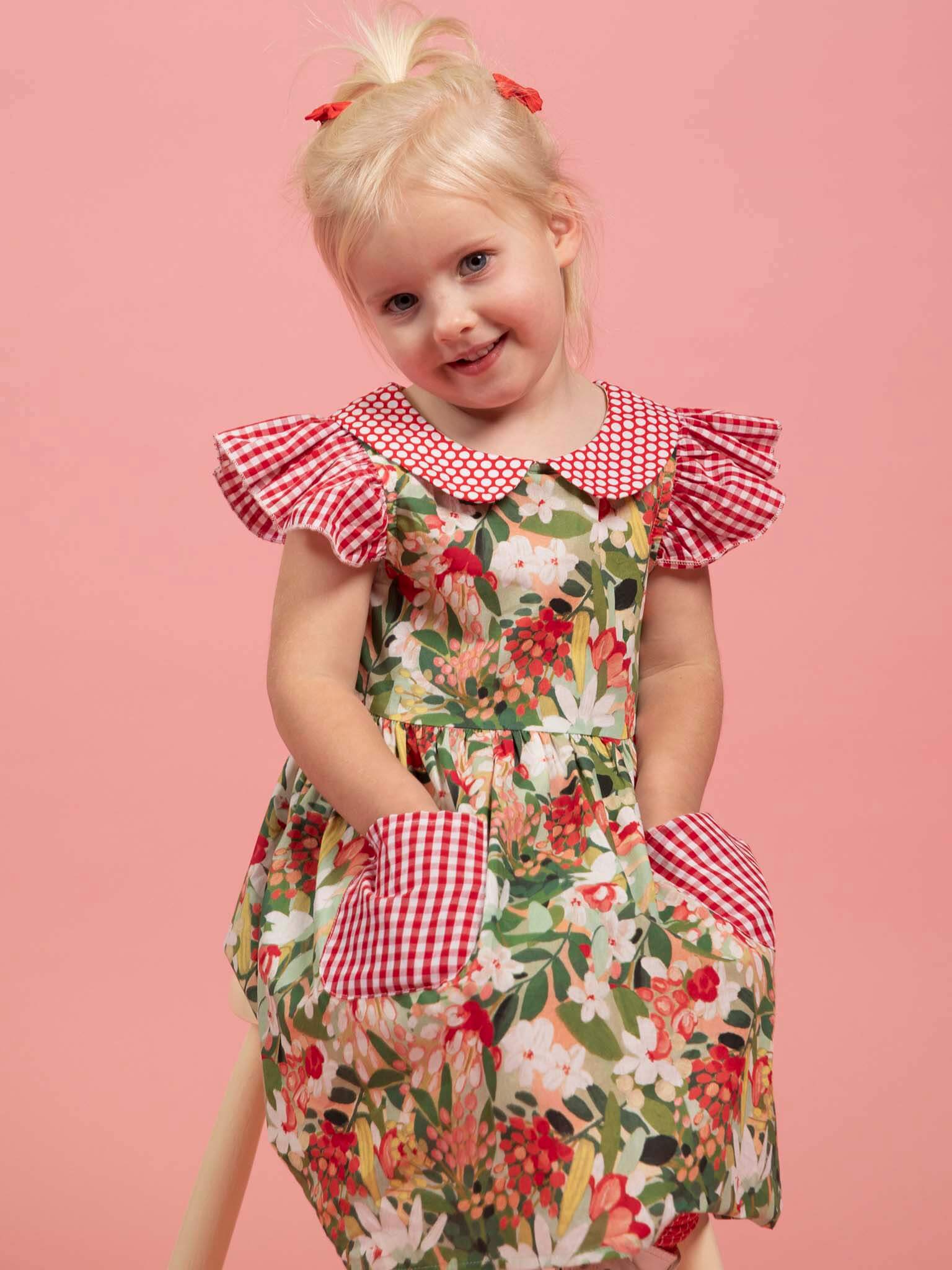 A young blonde-haired child poses against a pink background, wearing the Aussie Bush Flowers Collared Dress by Aussie Bush Flowers, featuring 100% cotton fabric with floral print and red gingham accents on the collar, sleeves, and pockets.