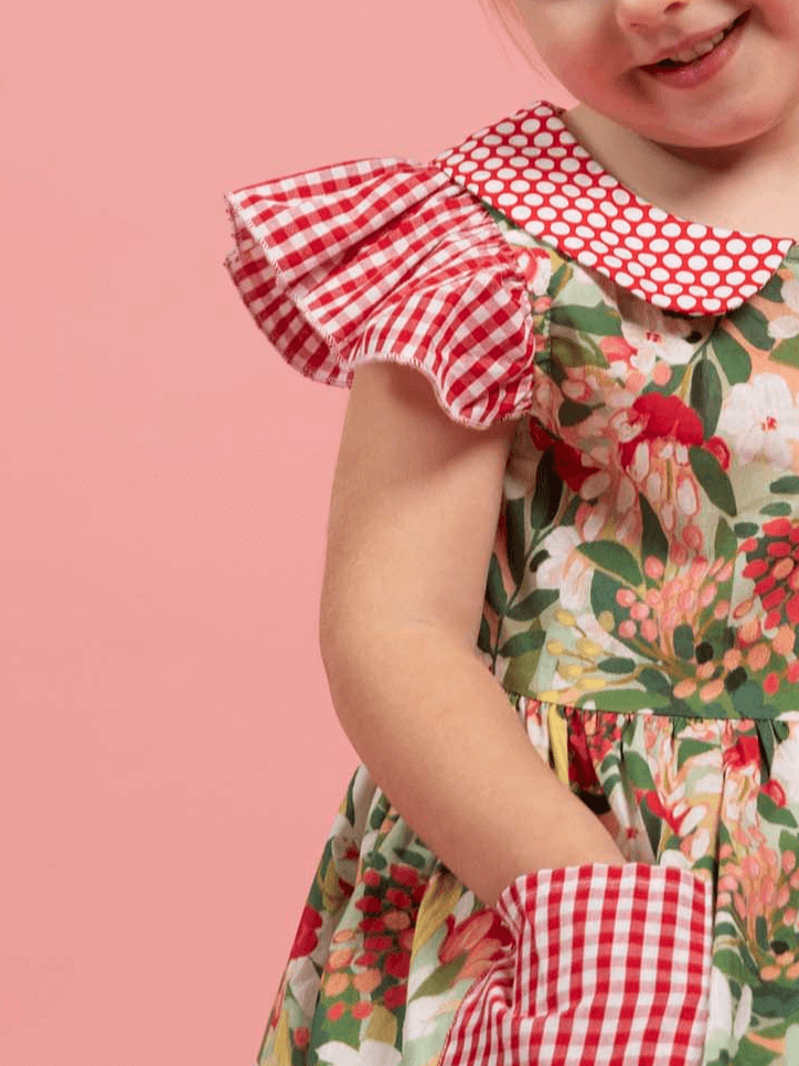 A young child in the Aussie Bush Flowers Collared Dress by Aussie Bush Flowers, featuring a floral print, red gingham ruffle sleeves, red polka dot collar, and 100% cotton fabric, stands against a pink background, partially smiling.