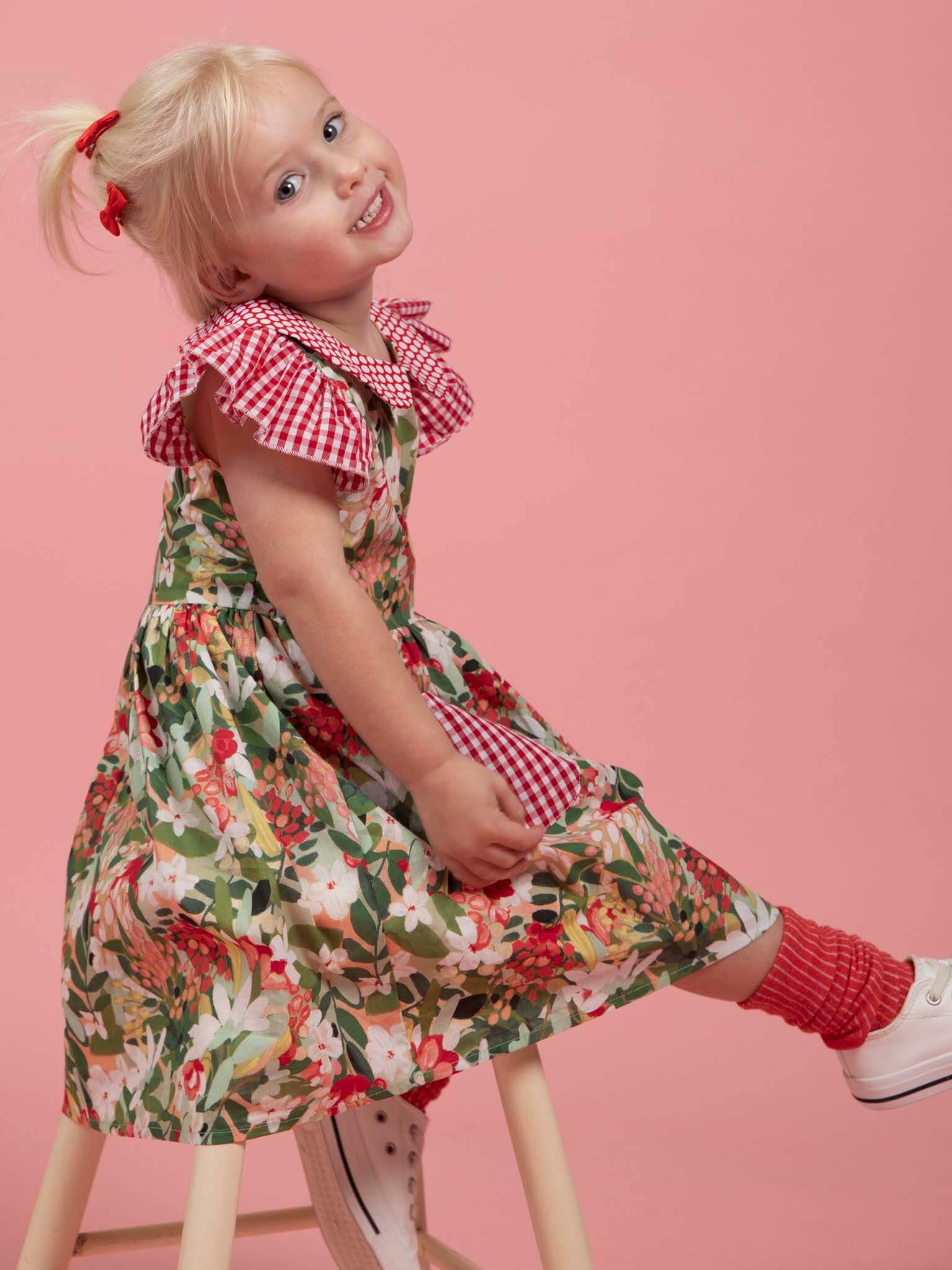 A young child with blonde hair in a ponytail wears the Aussie Bush Flowers Collared Dress by Aussie Bush Flowers, featuring floral and red checkered details, with red socks and white shoes, sitting sideways on a stool against a pink background.