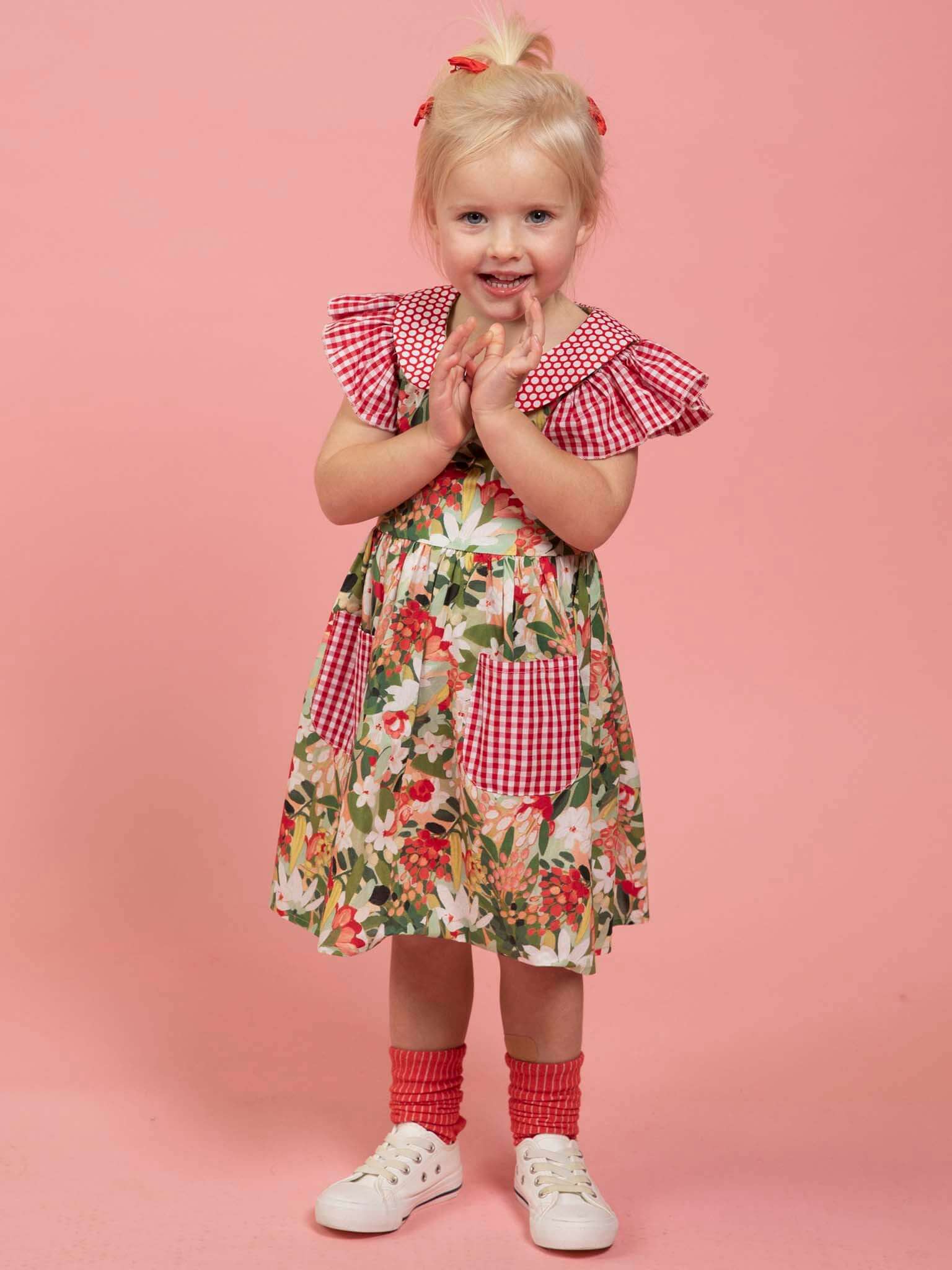 A young child with blonde hair wears the Aussie Bush Flowers Collared Dress by Aussie Bush Flowers, featuring a floral print with red gingham accents in 100% cotton, paired with red socks and white sneakers, standing against a pink background.