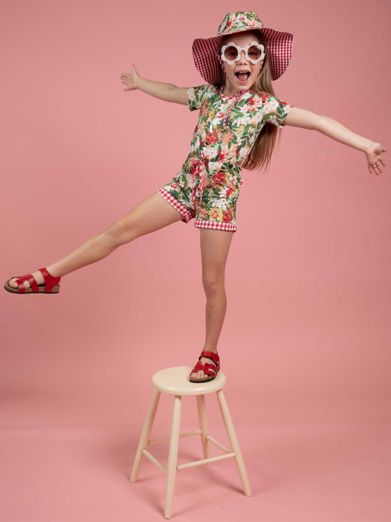 Child in floral outfit and sunglasses standing on a stool against a pink background