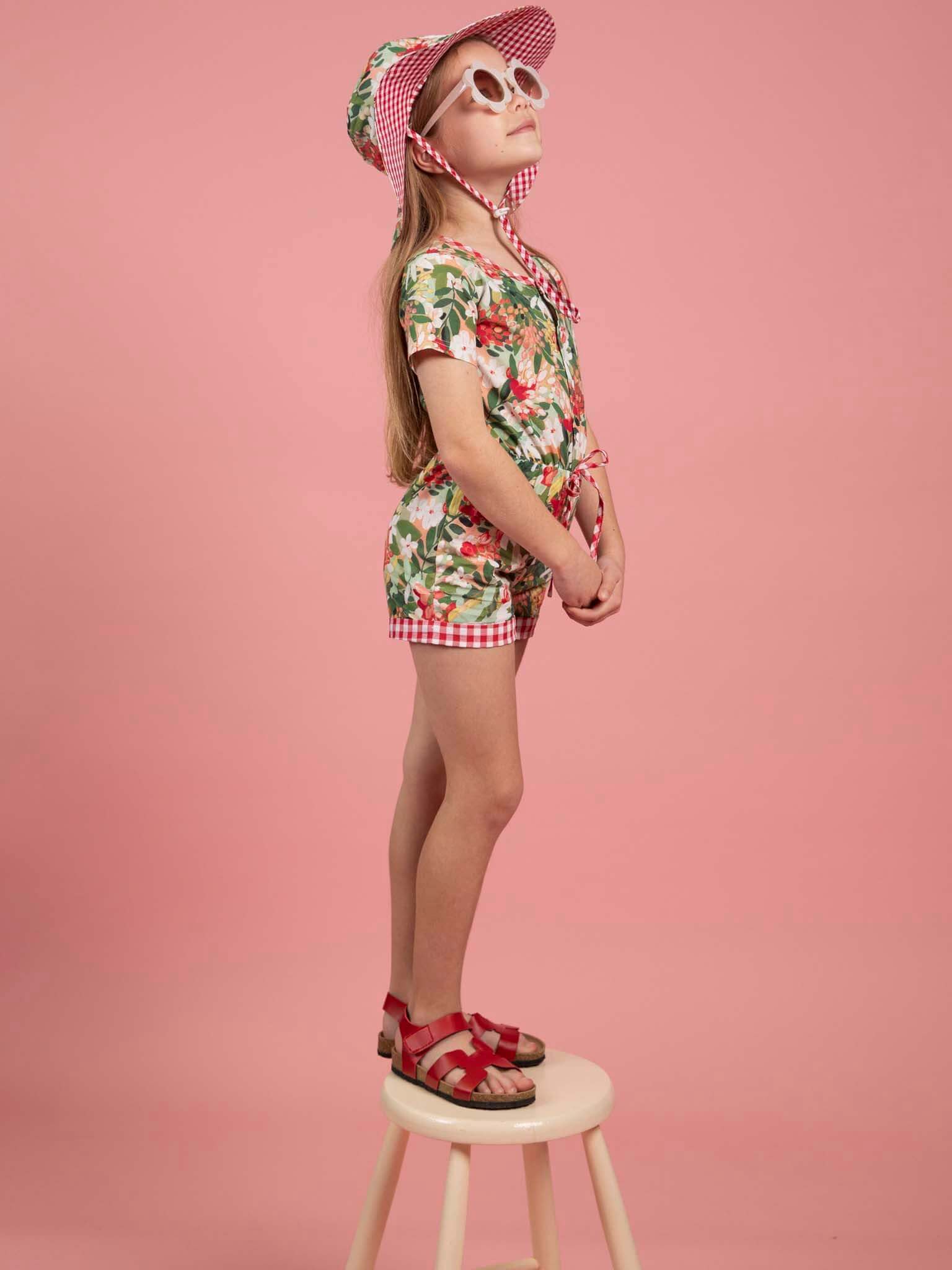 Young girl in a floral outfit and sun hat standing on a stool against a pink background