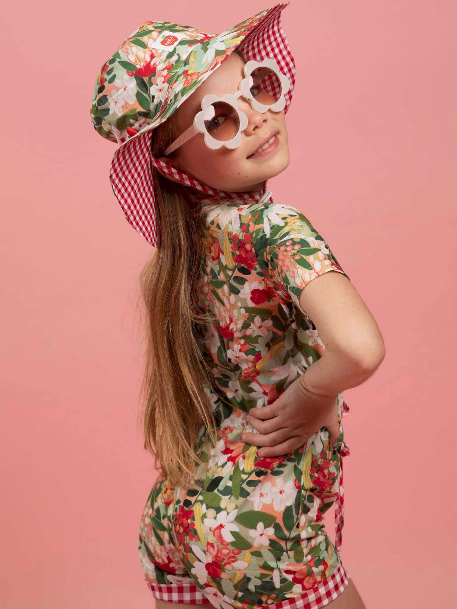Child wearing a floral outfit with a hat and sunglasses on a pink background