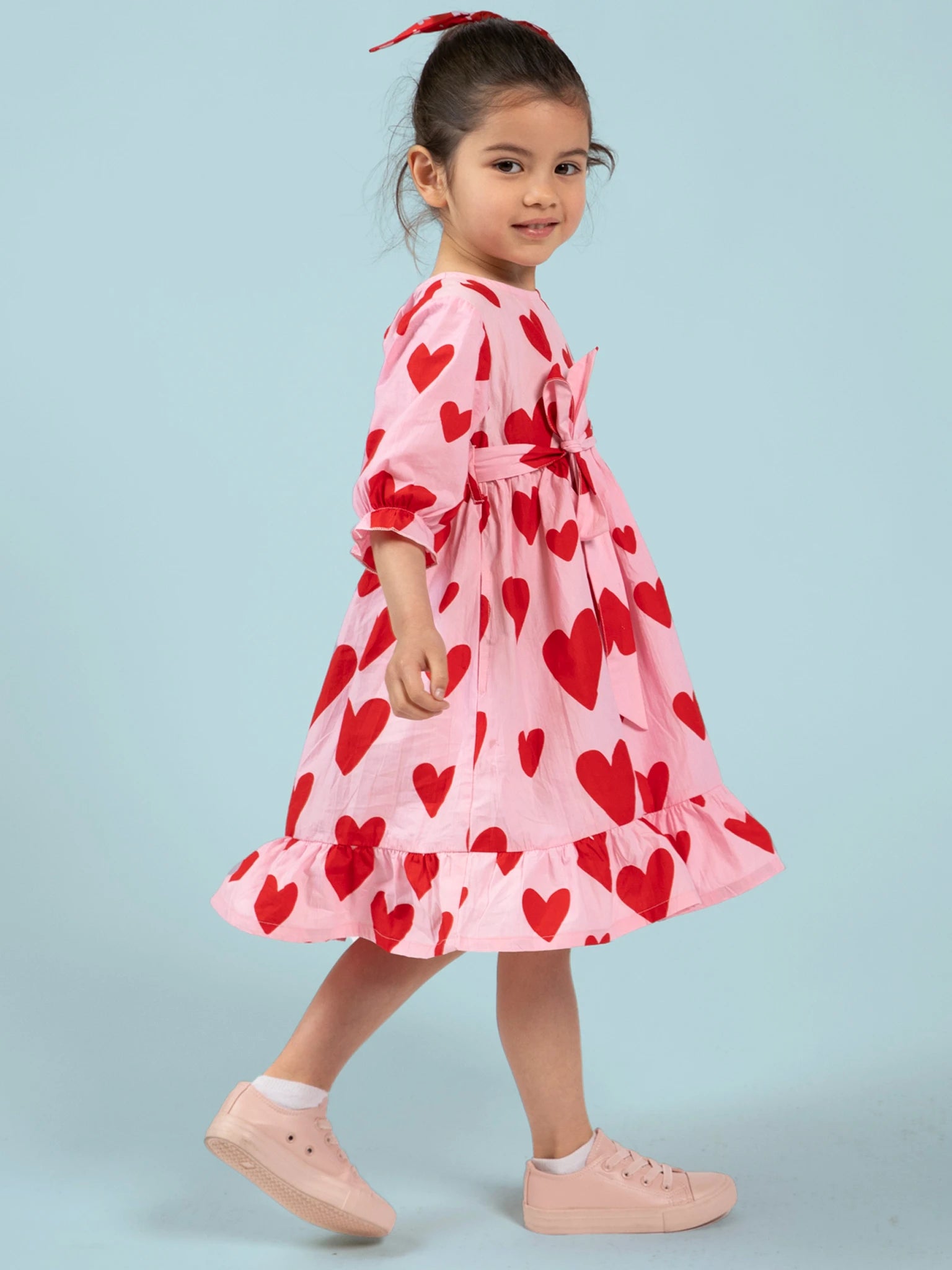 A young girl wearing the Big Hearts Big Heart Girl's Midi Dress—ethically made in India with handprinted red heart motifs—pairs it with pink sneakers and a red hairband, standing before a light blue backdrop.