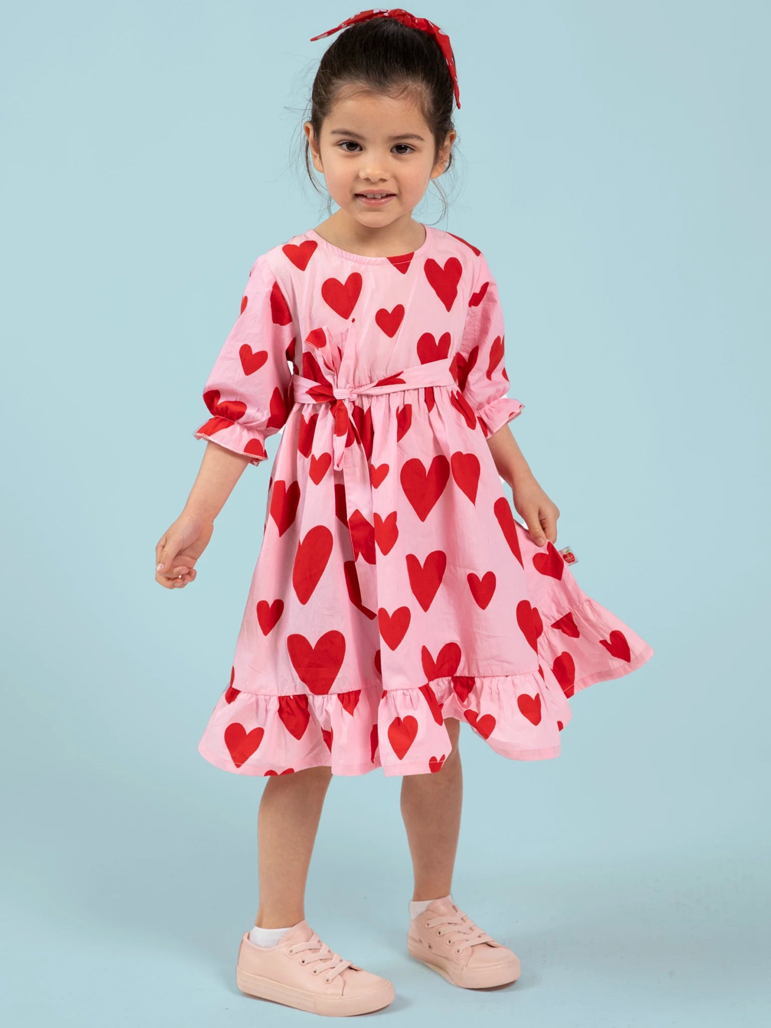 A young girl wears the Big Hearts Big Heart Girl's Midi Dress—ethically made in India—with red heart prints, paired with light pink sneakers and a red hair ribbon, standing against a light blue background.