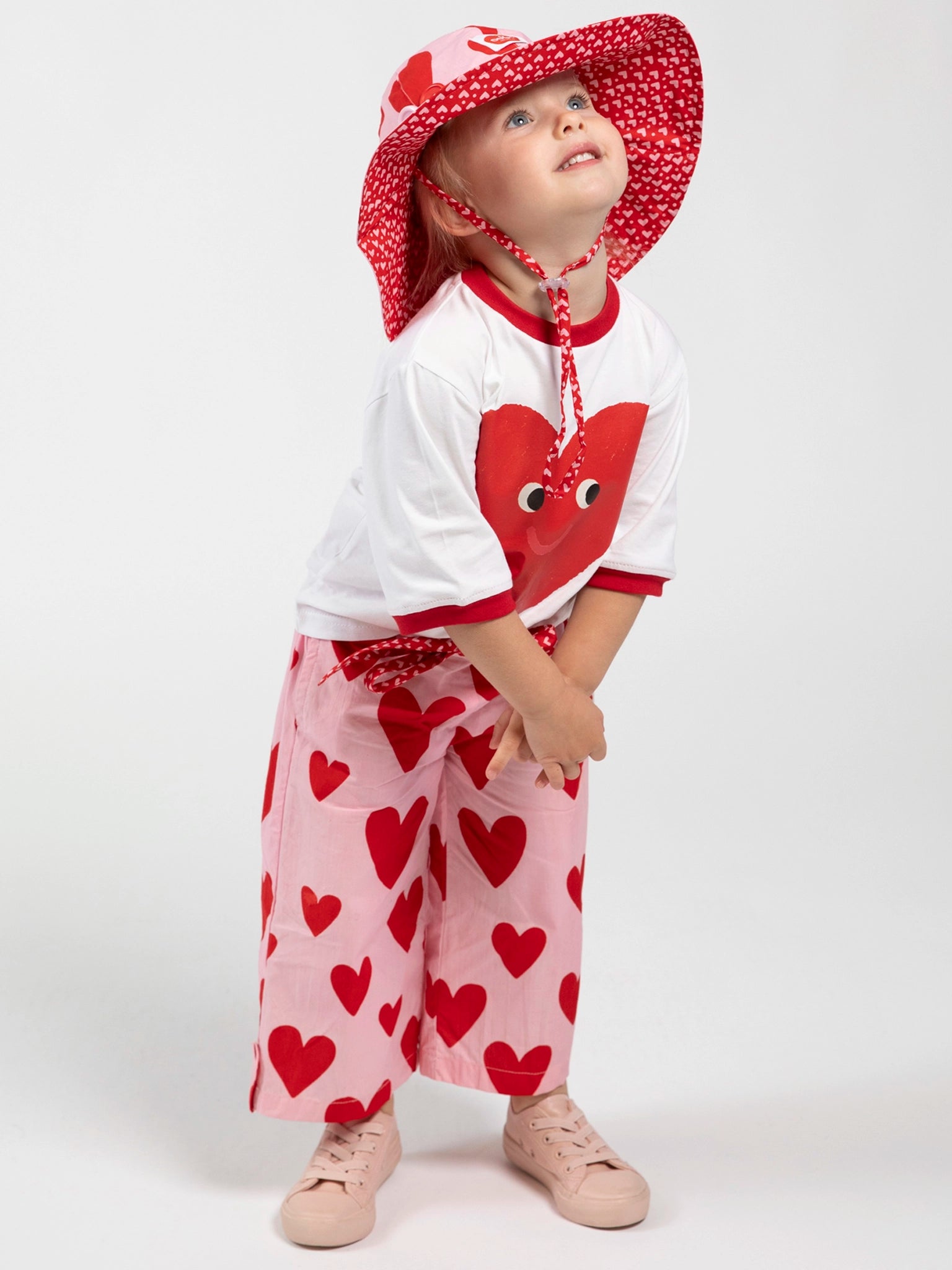 A young child in a white shirt with a red heart graphic, pink pants, and pink shoes wears the Big Hearts Big Heart Ponytail Hat with red hearts while standing against a plain white background.