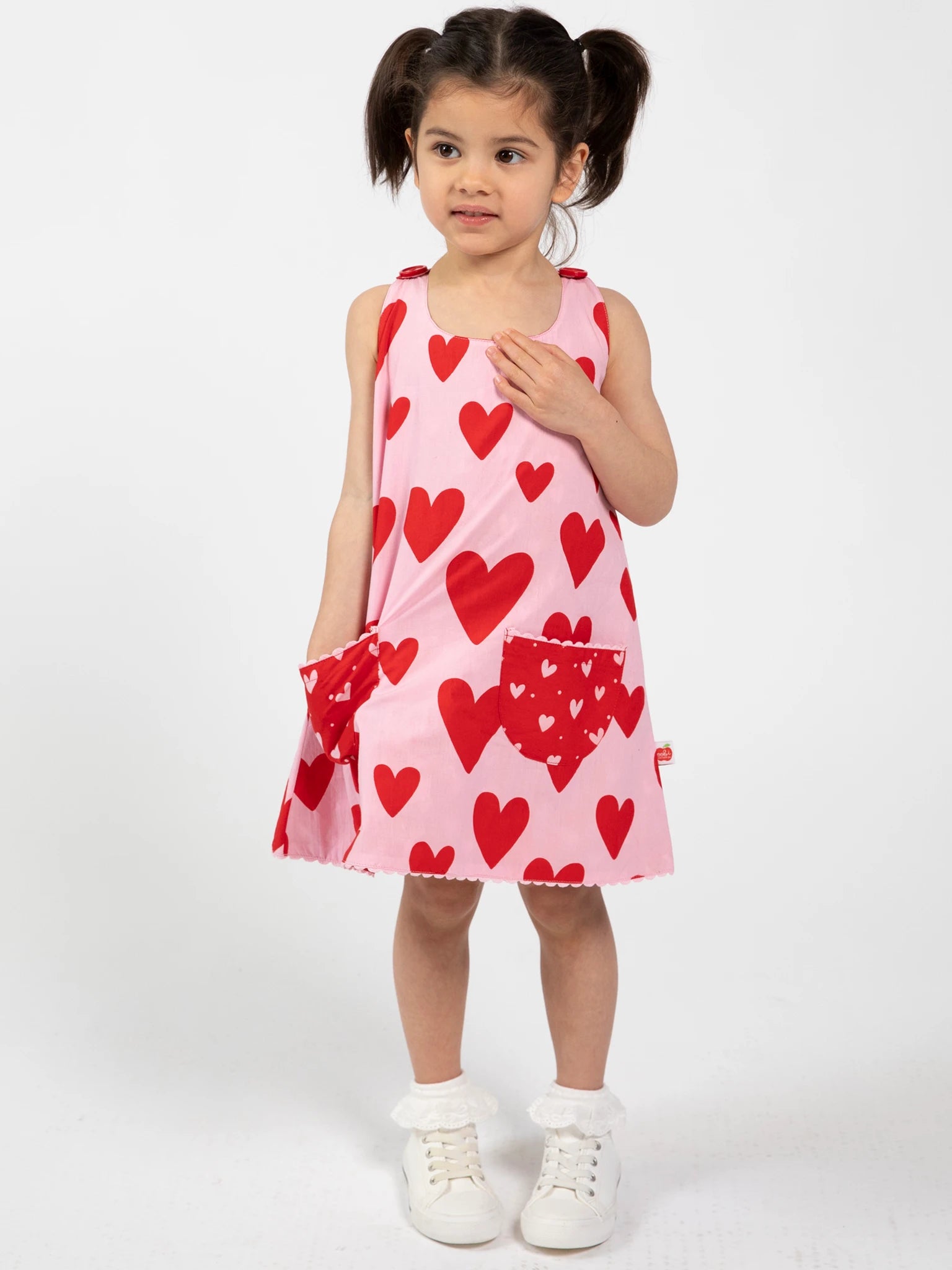 A young girl stands against a white background wearing the Big Hearts Big Heart Gracie Reversible Dress, featuring red heart patterns, paired with white socks and white sneakers.