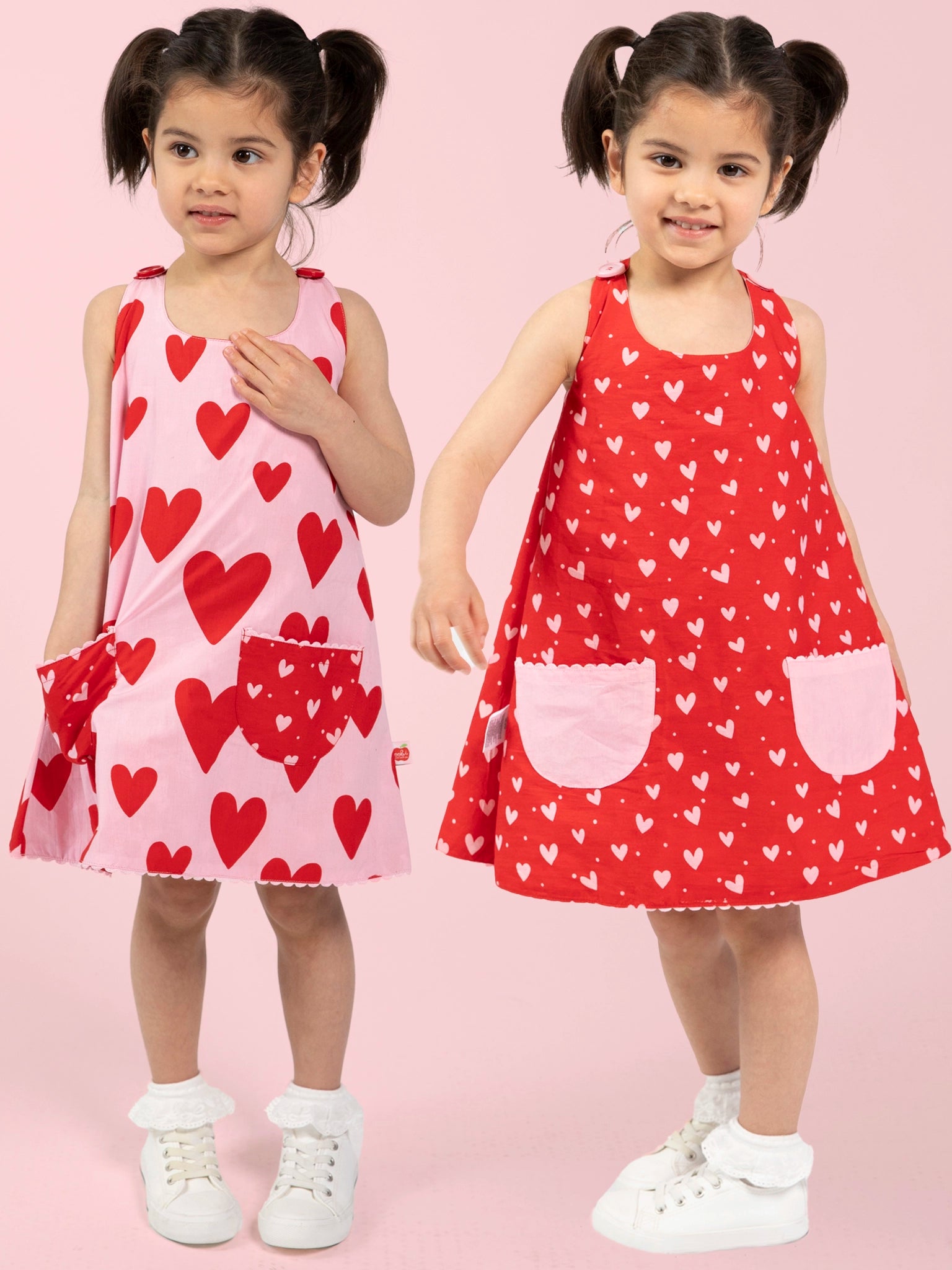 Two young girls with dark hair in pigtails wear Big Hearts’ Big Heart Gracie Reversible Dresses—one pink with red hearts, the other red with white hearts—standing against a pink background.