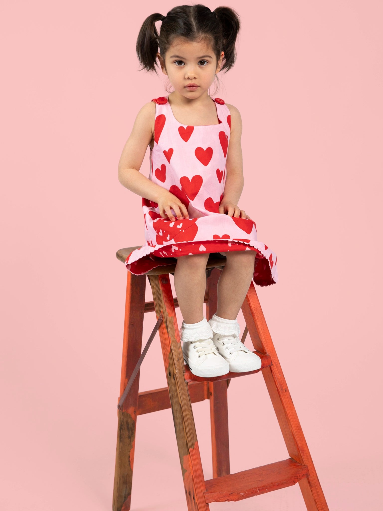 A young girl with pigtails sits on a wooden stepladder, wearing the Big Hearts Big Heart Gracie Reversible Dress against a pink background, paired with white socks and shoes.