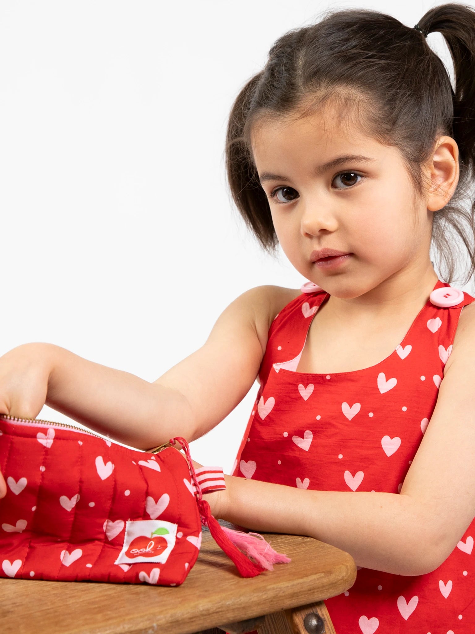 A young girl in a red, heart-patterned dress sits at a wooden table, reaching into an Oobi Heart Print Quilted Pouch featuring heart designs and a shimmering gold zipper.