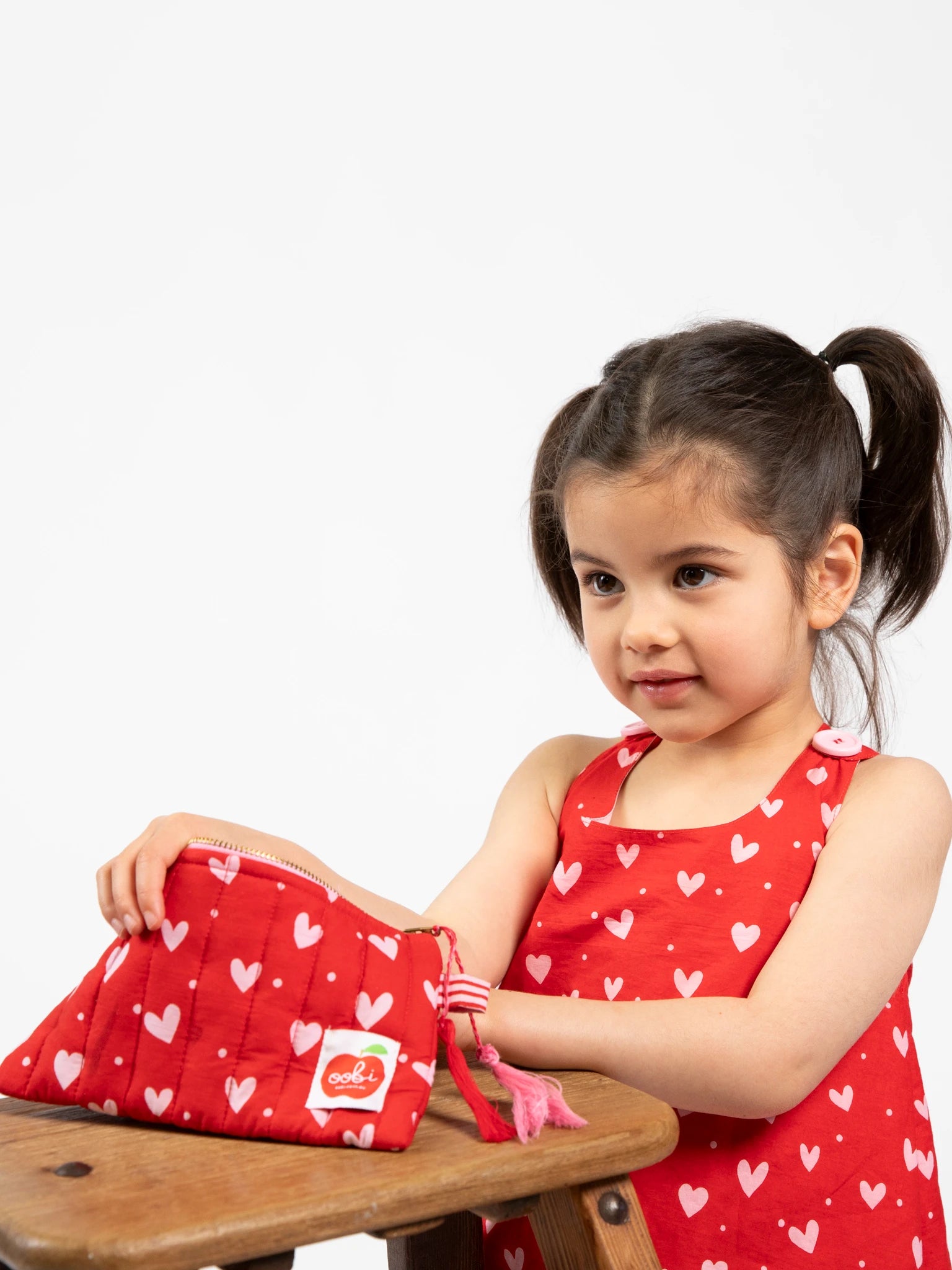 A young girl in a red dress with white hearts sits at a wooden table, holding the Oobi Heart Print Quilted Pouch decorated with handmade tassels and white heart patterns.