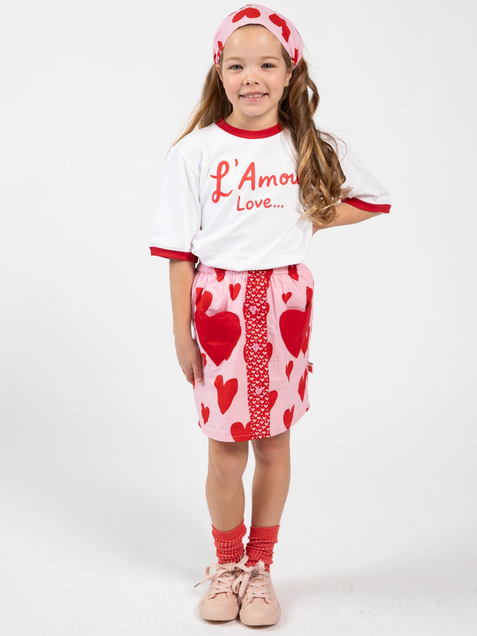 A young girl smiles in a white "L'Amour Love..." tee, Big Hearts' Big Heart Pocket Skirt with red hearts and heart-shaped pockets, matching socks, pink shoes, and a heart-print headband.