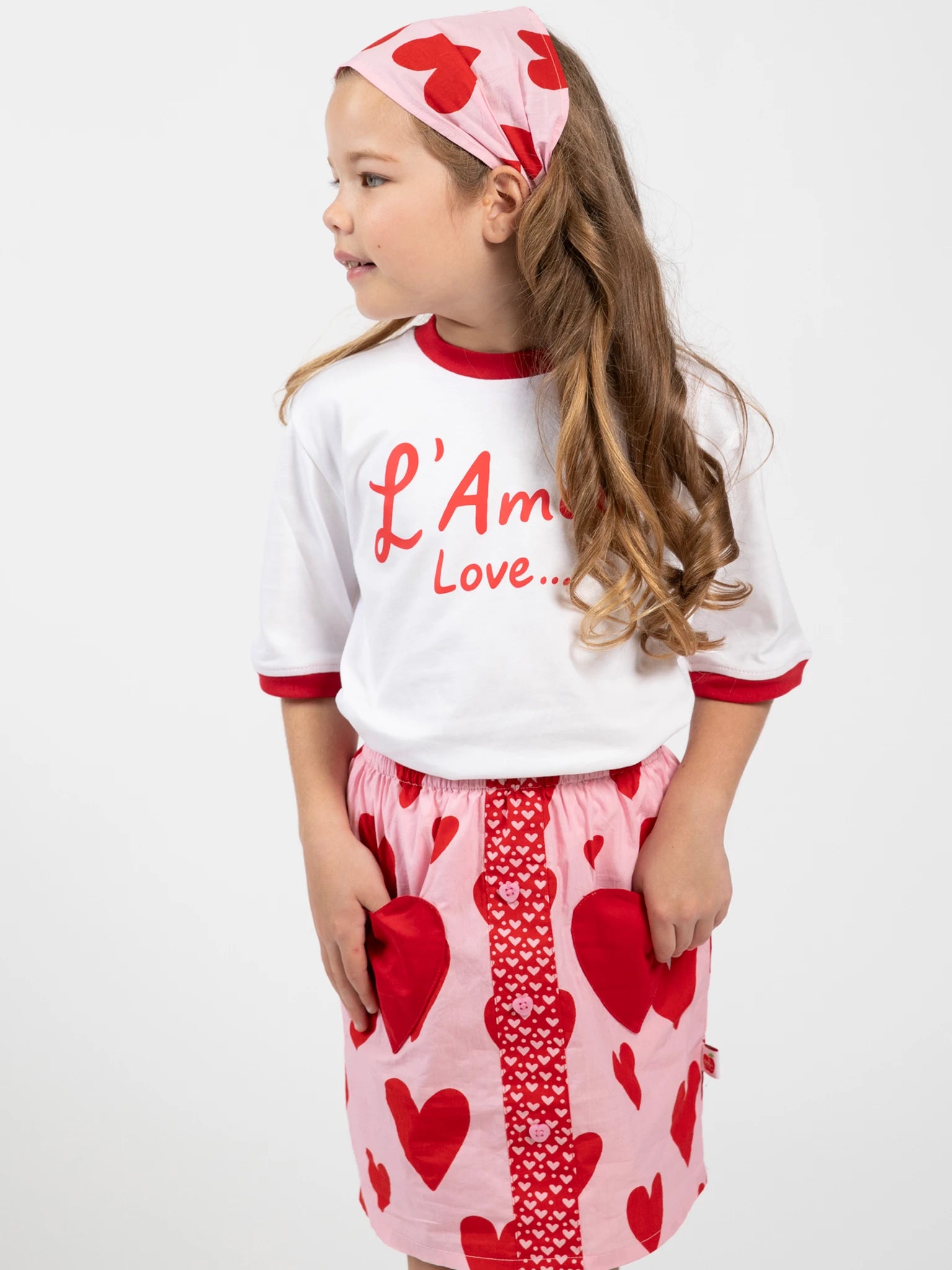 A young girl models the Big Hearts Big Heart Pocket Skirt—pink, hand-printed cotton with heart-shaped pockets—paired with a white shirt, a matching headband, and set against a plain white background.