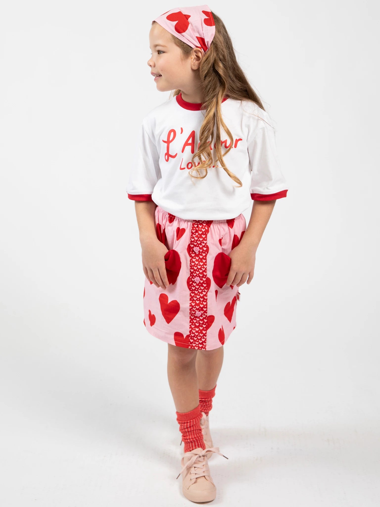 A young girl wears the Big Hearts Big Heart Pocket Skirt with heart-shaped pockets, paired with a white shirt with red trim, matching headband, red socks, and pink shoes, standing against a plain white background.