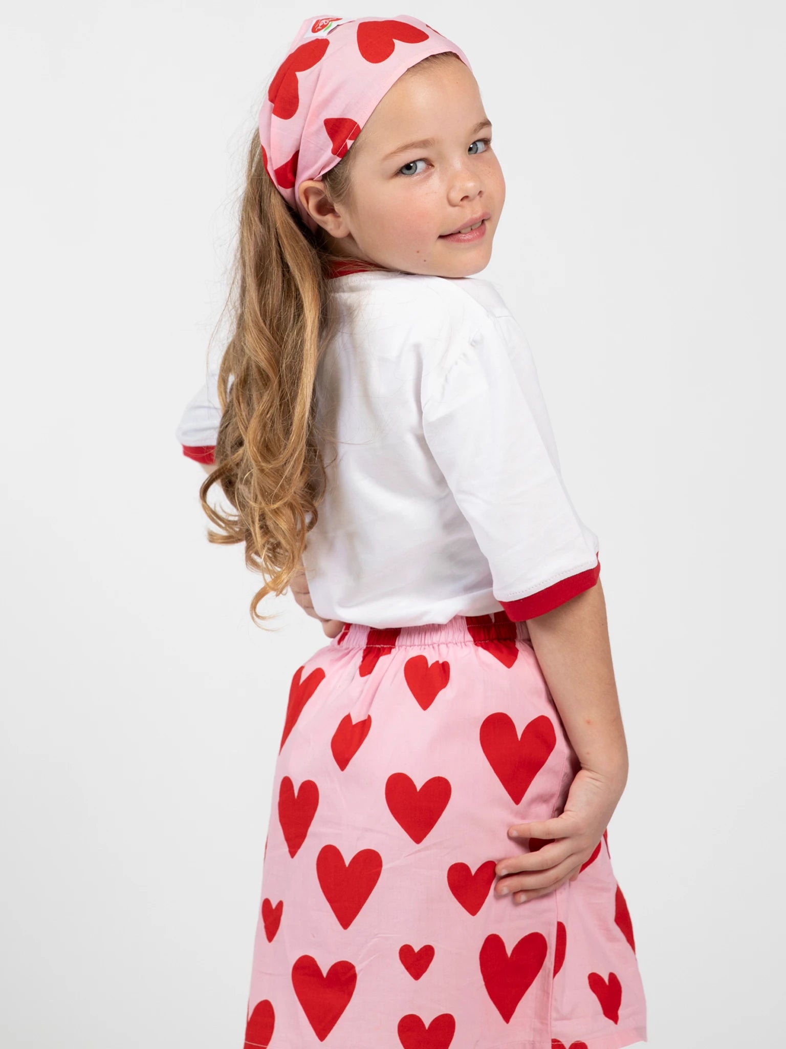 A young girl with long hair wears the Big Hearts Big Heart Pocket Skirt with heart-shaped pockets, a white shirt, and a matching headband, posing over her shoulder against a plain background.