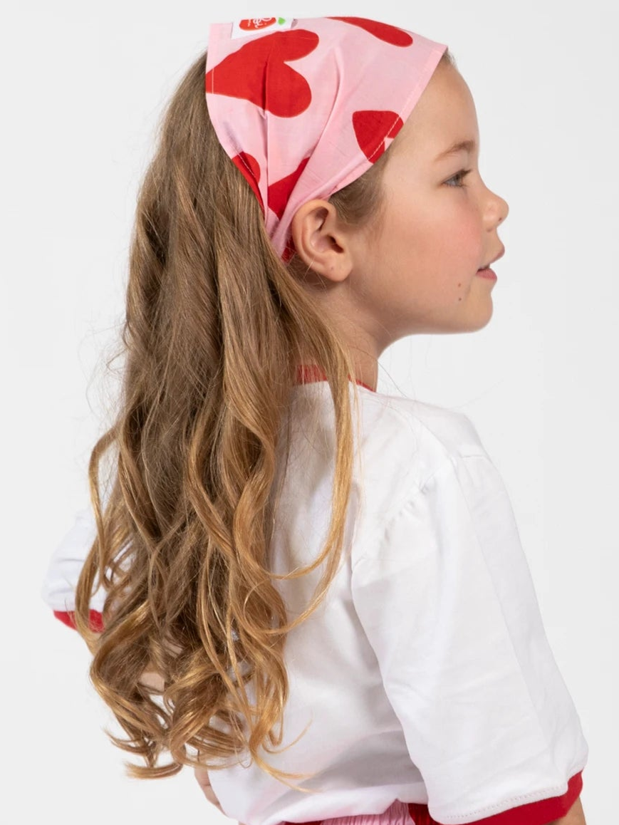 A young girl in profile wears the Essentials Big Heart Headband, featuring a hand-printed hearts pattern, styled with a white shirt and her long wavy hair draped over her shoulder for a retro look.