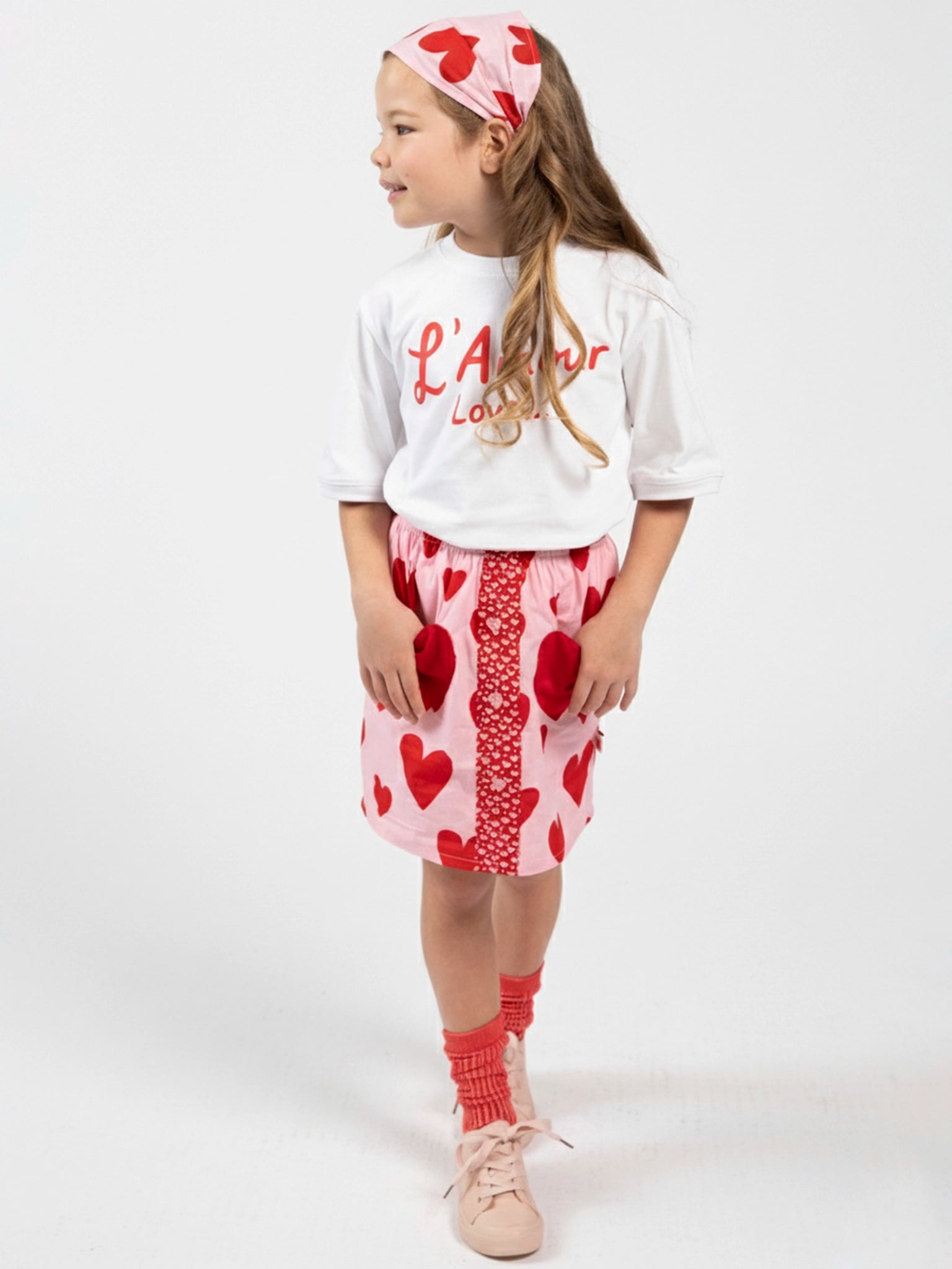 A young girl wears the Essentials L'Amour (Love) Tee with Red Binding (100% cotton), paired with a pink heart skirt, matching headband, red socks, and light pink shoes, standing against a white background.